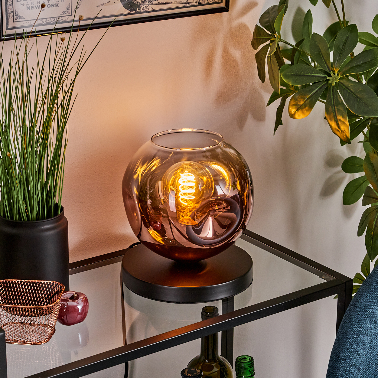 Eine dekorative Lampe mit Glühbirne steht auf einem Glasregal. Links ist eine Pflanze, ein Zierapfel und ein Korb.