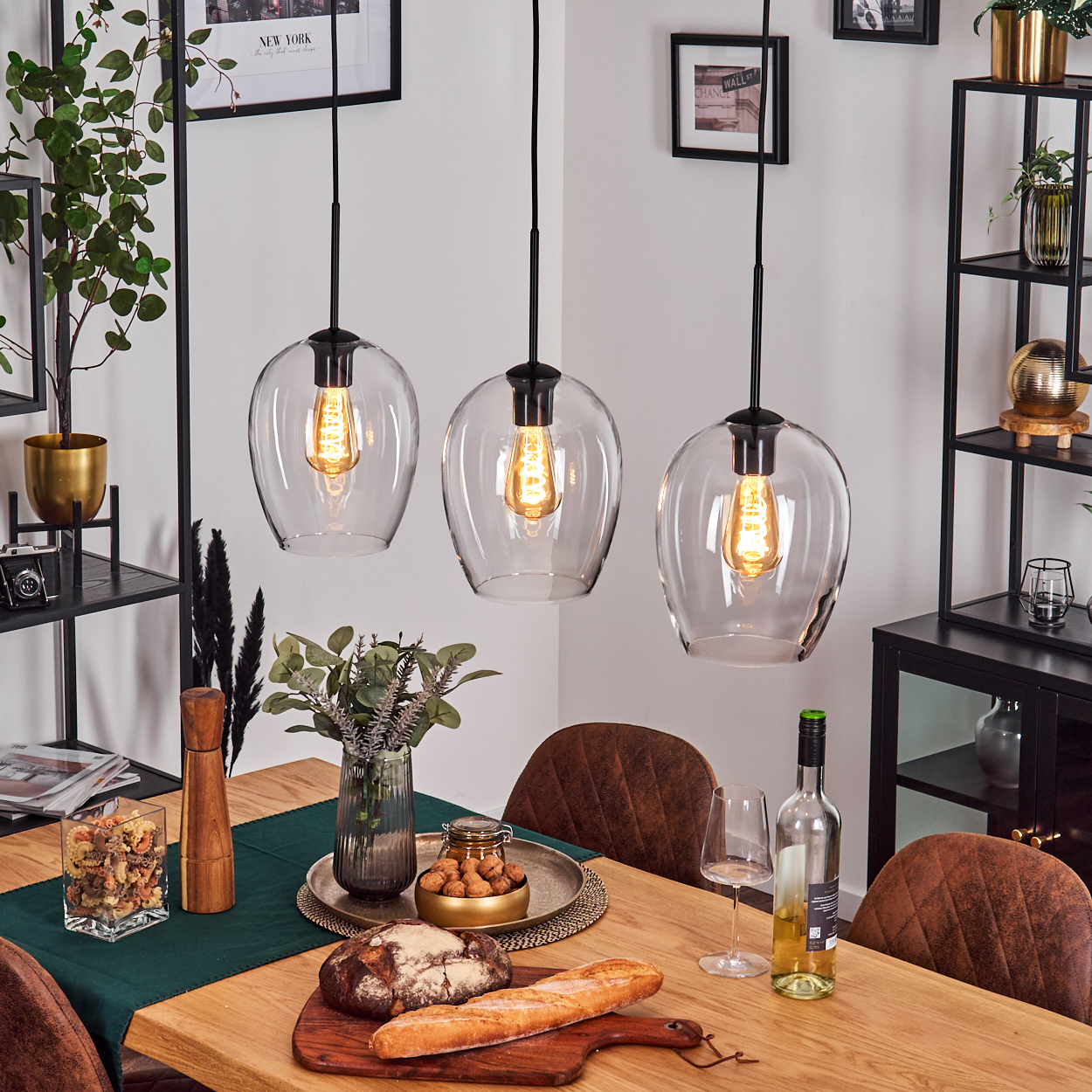 Esszimmer mit drei Hängelampen. Holztisch mit Essen und Wein. Stühle mit braunem Leder. Regale mit Pflanzen.
