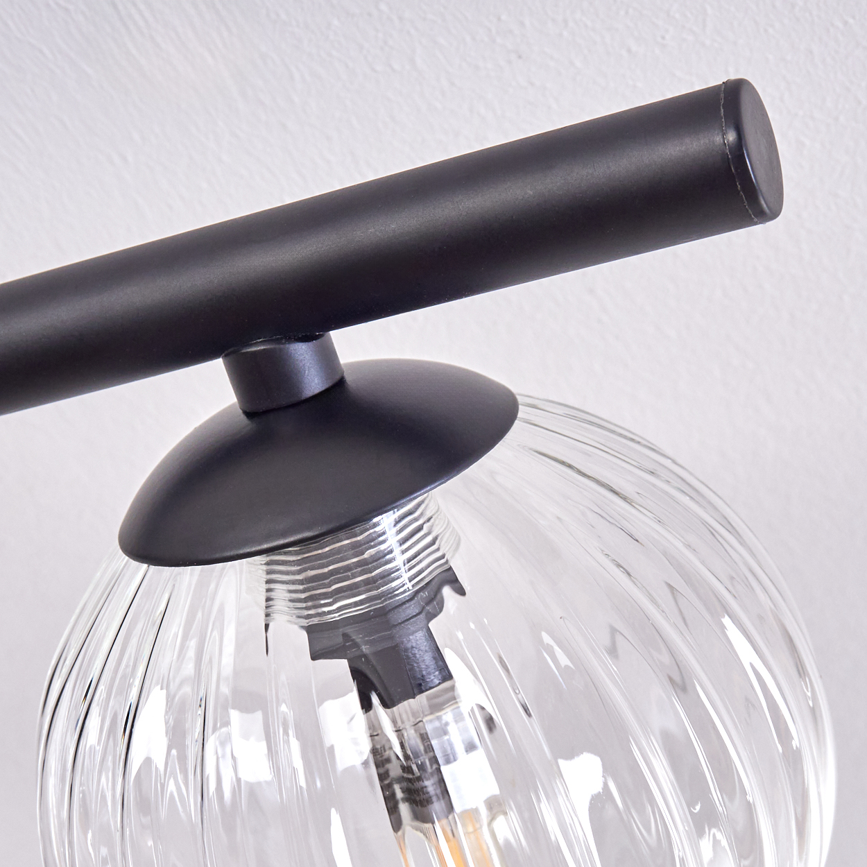 Nahaufnahme einer schwarzen Lampe mit einem klaren, geriffelten Glaskolben. Der Hintergrund ist eine schlichte, weiße Wand.