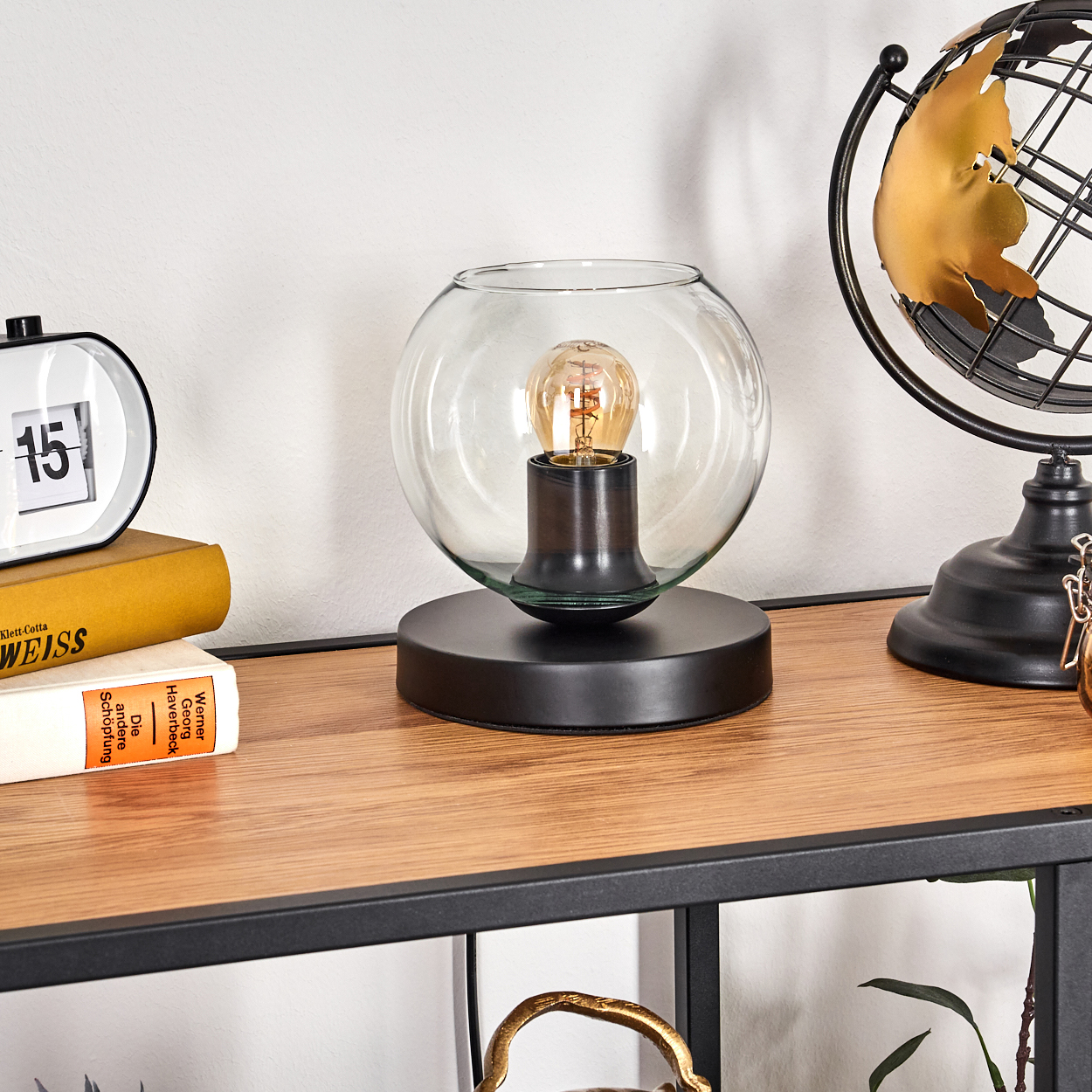 Ein Schreibtisch mit Lampe, Globus, Uhr und Büchern. Die Lampe hat eine Glühbirne in einer Glaskugel. Der Globus ist goldfarben.