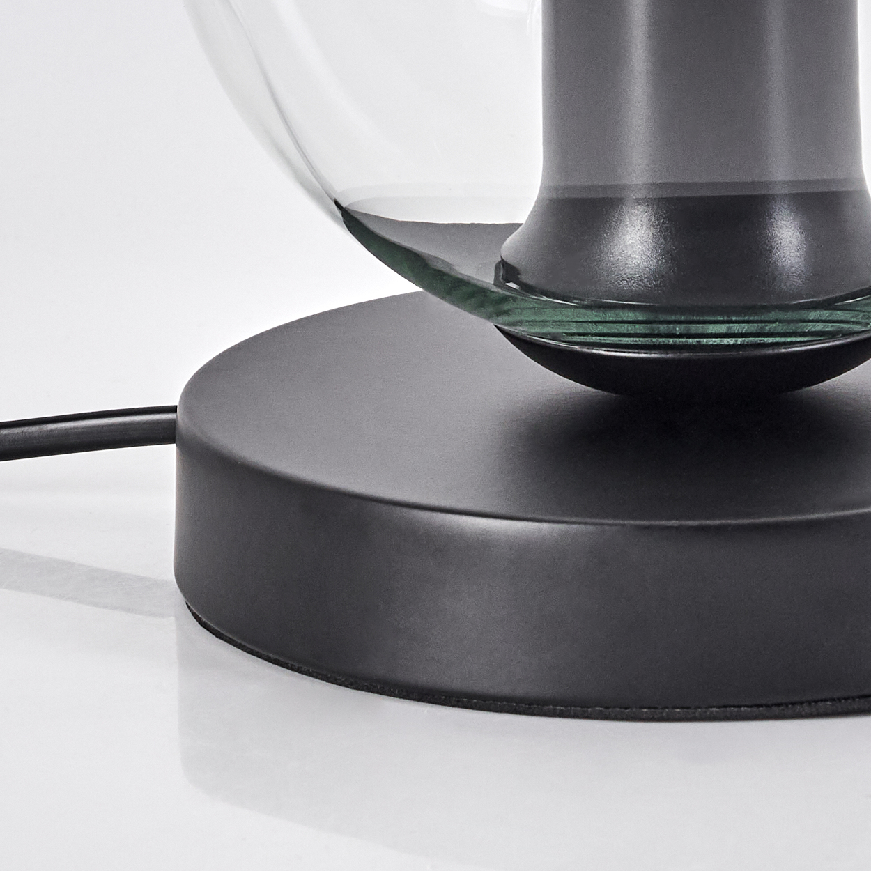 Nahaufnahme einer modernen Lampe. Sie hat einen schwarzen Sockel, einen Glasschirm und ein schwarzes Kabel. Die Lampe steht auf einer weißen Oberfläche.