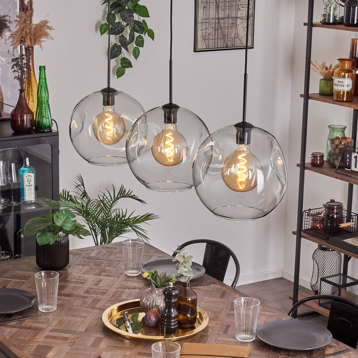 Esszimmer mit Holztisch, drei Glaskronleuchtern und dekorativen Gegenständen. Pflanzen und Gläser stehen auf dem Tisch.