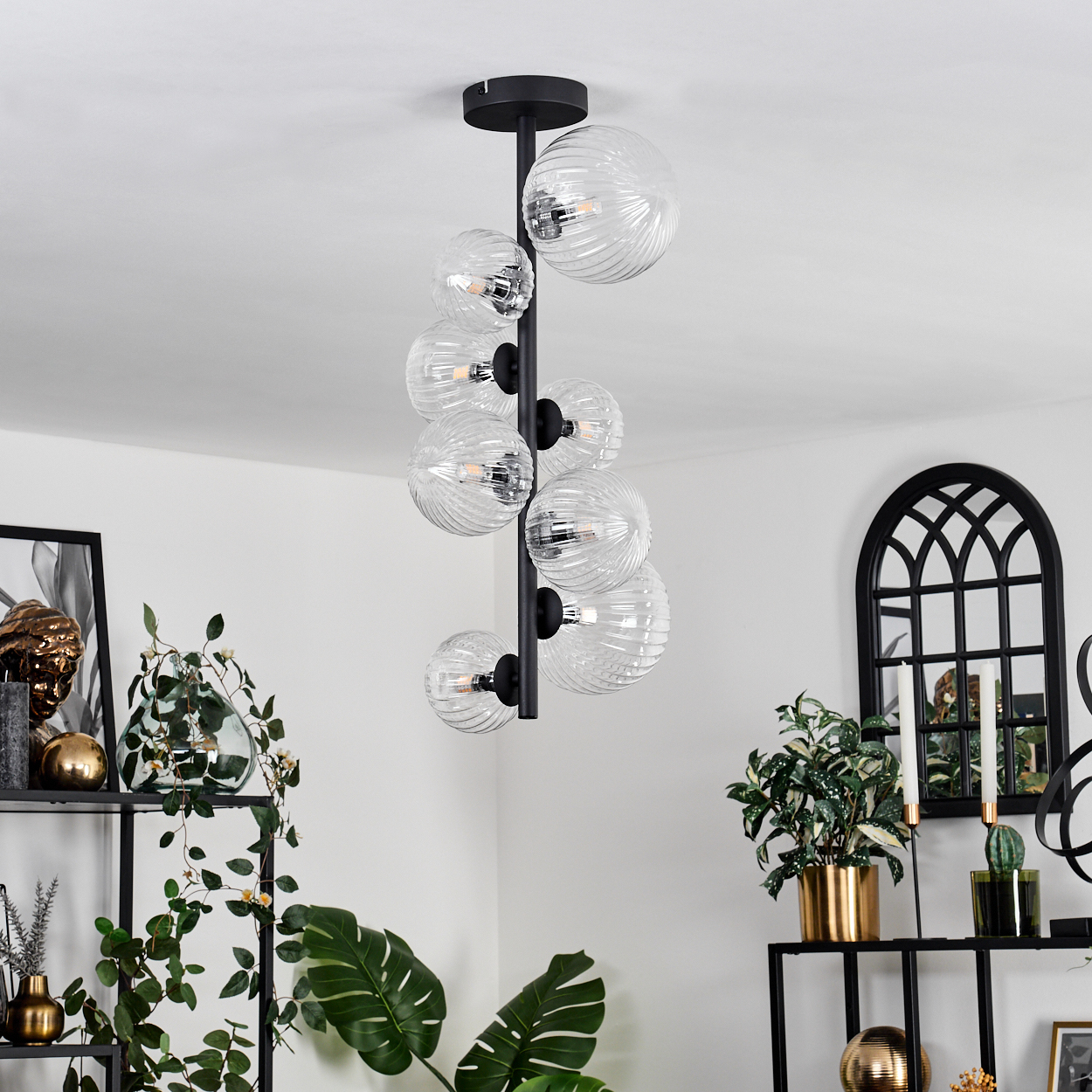 Ein moderner Kronleuchter mit Glaskugeln hängt in einem Raum mit Pflanzen und Dekoration. Schwarze Metallregale und ein Fensterrahmen sind sichtbar.