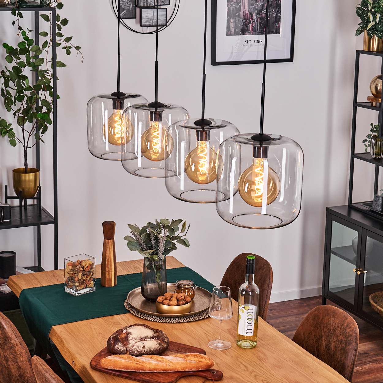 Esszimmer mit Holztisch. Hängelampen, Essen, Wein und Stühle sind sichtbar. Regale mit Pflanzen und Dekoration sind vorhanden.