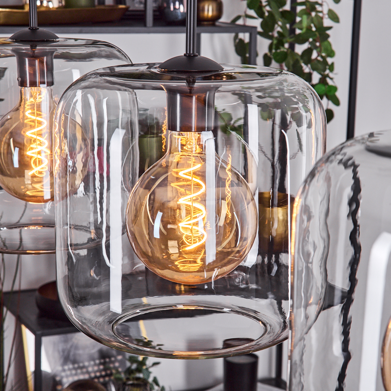 Nahaufnahme von Glas-Pendelleuchten mit glühenden Wendelglühlampen. Klare Glasschirme reflektieren Licht. Der Hintergrund umfasst Deko und Pflanzen.