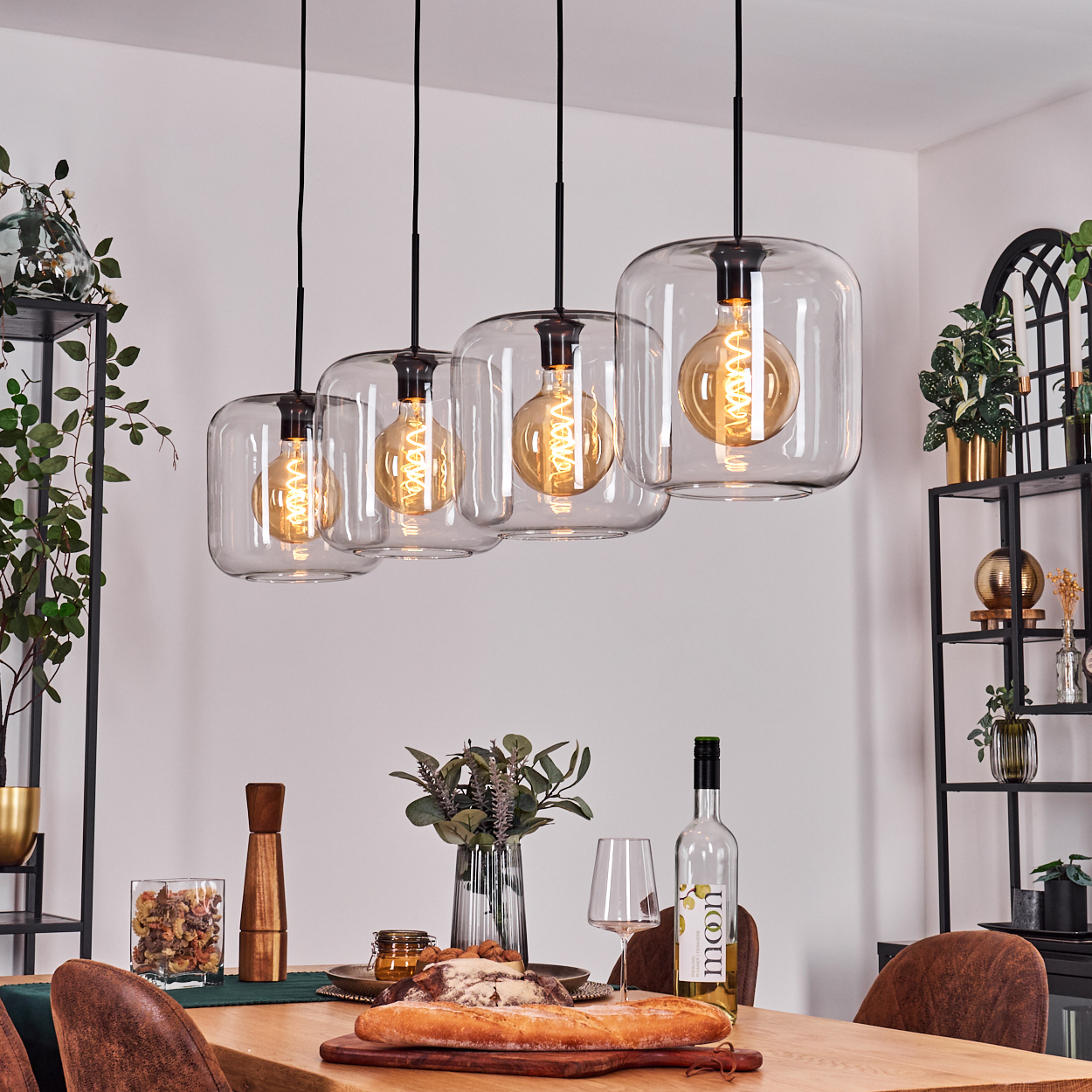 Esszimmer mit Holztisch. Vier Hängelampen mit sichtbaren Glühbirnen. Brot, Wein und Pflanzen stehen auf dem Tisch.