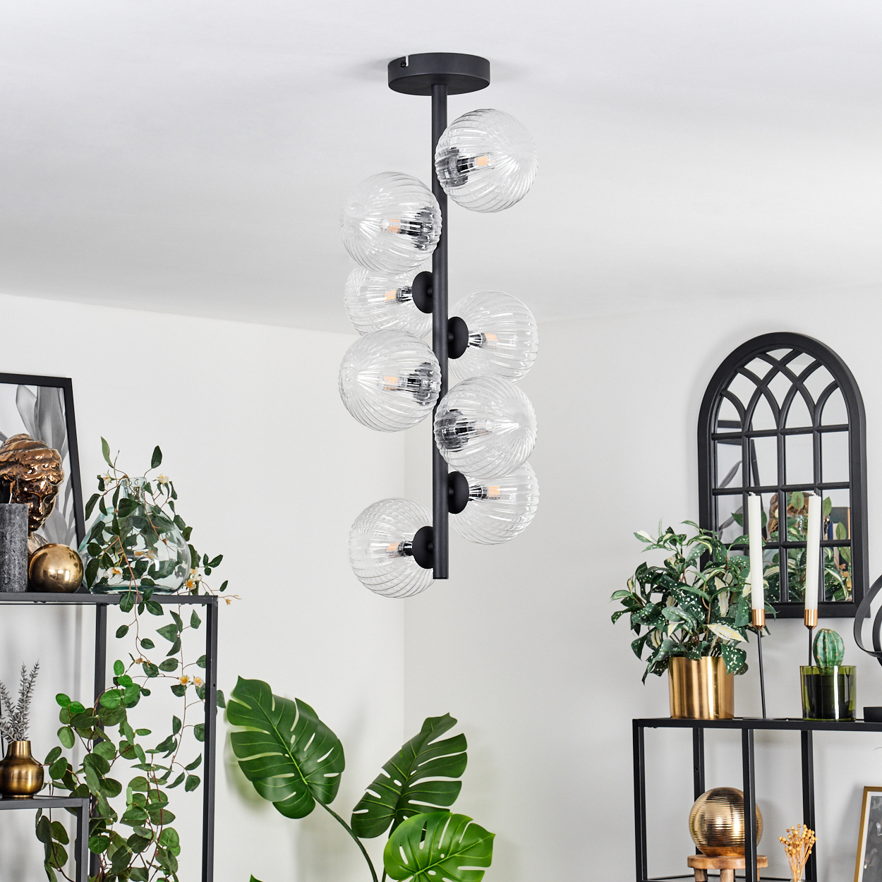 Ein moderner Kronleuchter mit Glaskugeln hängt in einem Raum. Pflanzen und dekorative Gegenstände stehen auf Regalen. Ein Spiegel ist an der Wand.