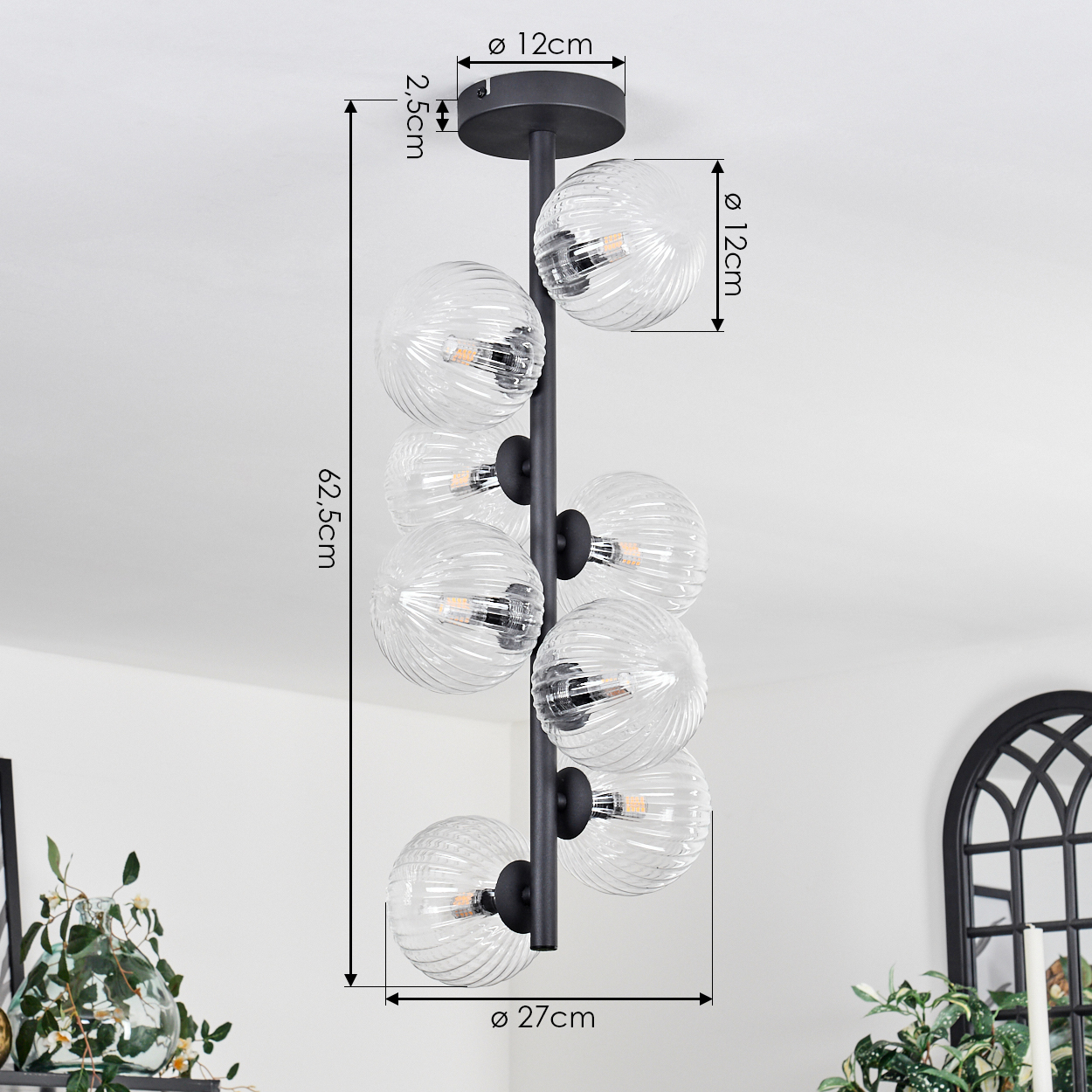 Eine moderne Deckenleuchte mit Glasschirmkugeln hängt von der Decke. Abmessungen sind beschriftet. Der Hintergrund ist eine weiße Wand mit Dekoration.