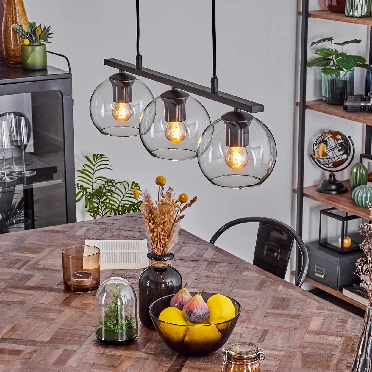 Esstisch mit Dekoration, darunter eine Glashängeleuchte. Auf dem Tisch sind Früchte, Blumen und Kerzen. Regale mit Dekoration im Hintergrund.