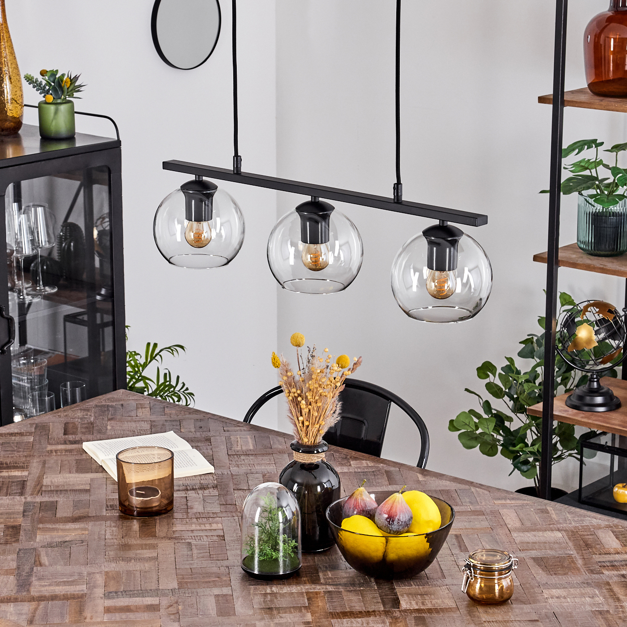 Esszimmer mit Holztisch, Glaslampen und einer Obstschale. Schwarze Regale, Schrank und Spiegel sind sichtbar.