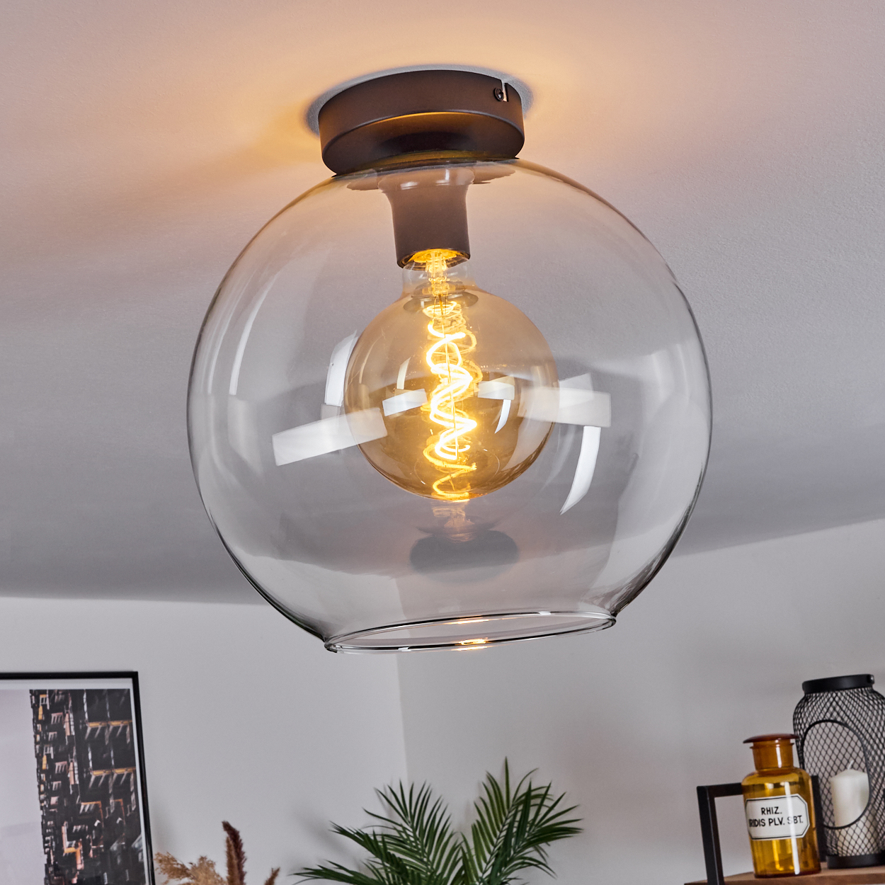 Eine klare Glaskugel-Leuchte hängt von der Decke. Innen ist eine leuchtende Wendelbirne sichtbar. Im Hintergrund ist andere Dekoration zu sehen.