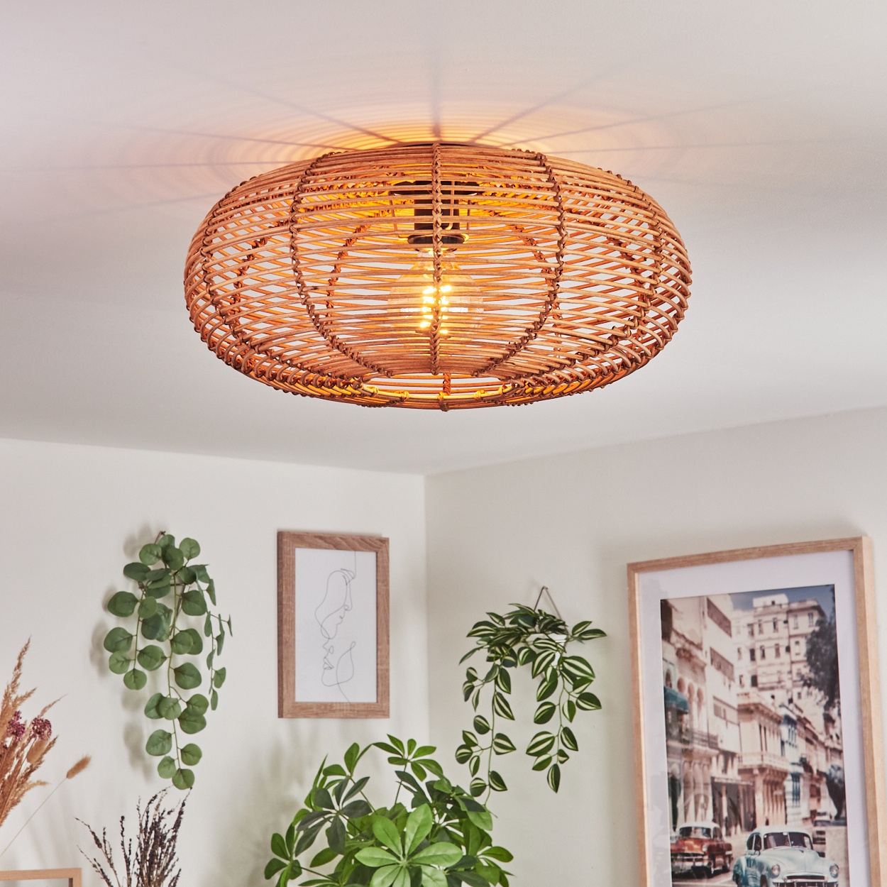 Eine gewebte Deckenlampe aus Rattan beleuchtet einen Raum mit Pflanzen und gerahmten Bildern. Die Lampe ist der Mittelpunkt, mit warmem Licht.