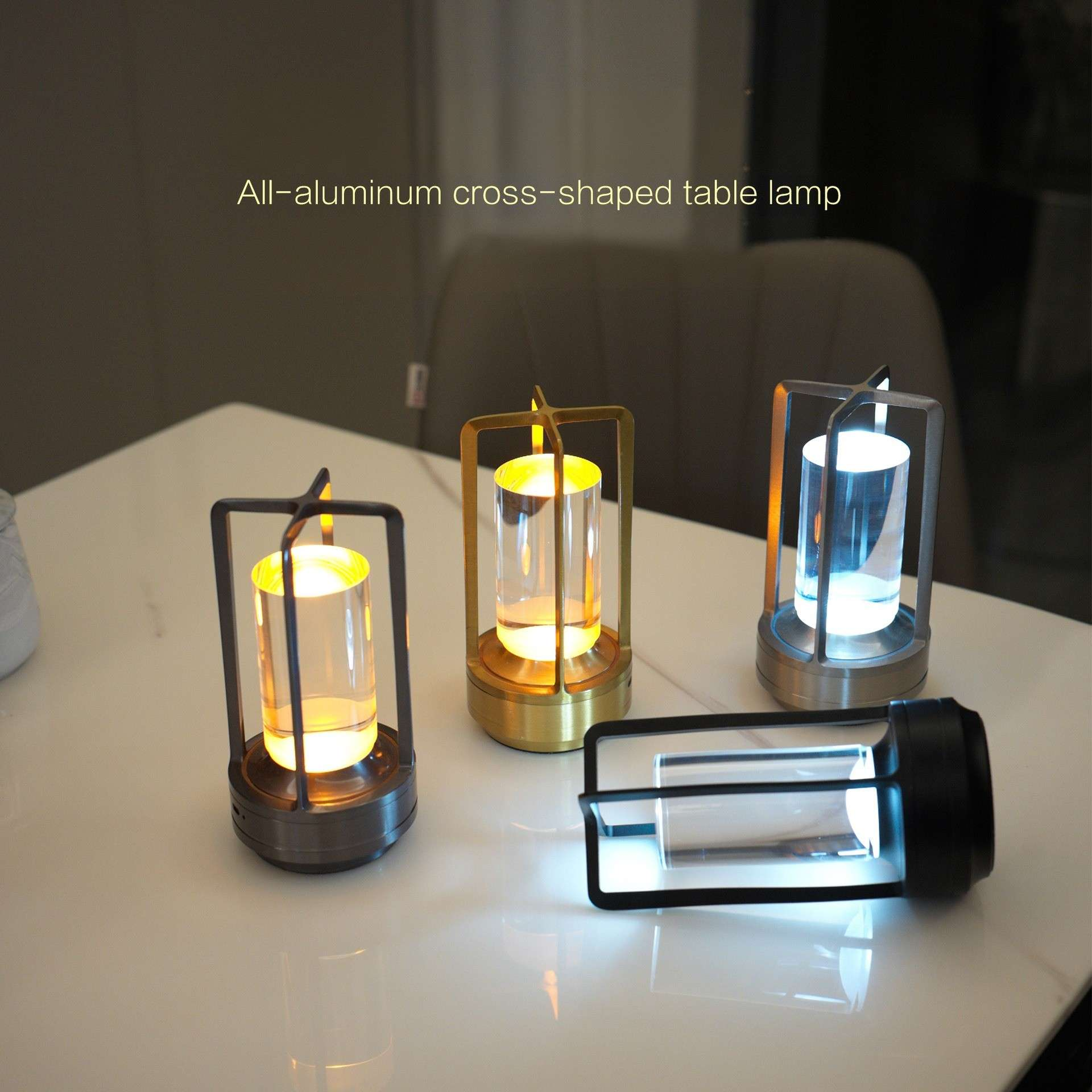 Vier kreuzförmige Tischlampen auf einer weißen Oberfläche. Sie haben zylindrische Lichtquellen in Metallrahmen und -sockeln.