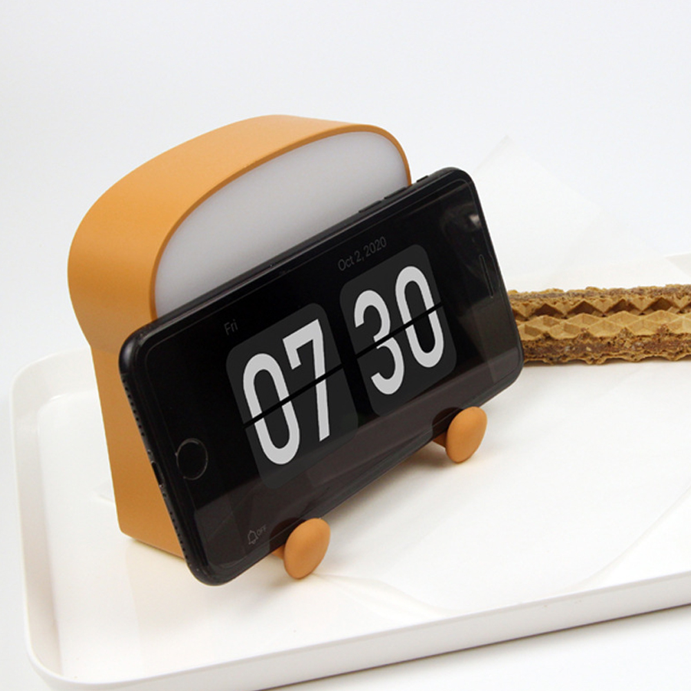 Ein orangefarbener, brotförmiger Telefonhalter mit einem Telefon, das 07:30 Uhr anzeigt. Ein Waffelkeks liegt auf einem weißen Tablett.