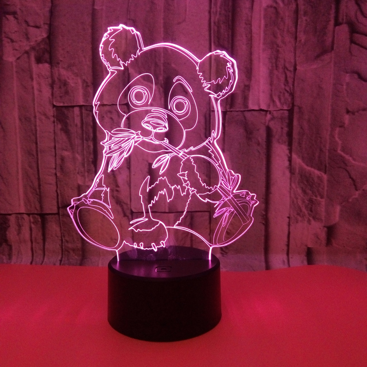 Eine pandaförmige LED-Lampe leuchtet rosa. Sie steht auf einem schwarzen Sockel, mit einer rosafarbenen Oberfläche darunter und einer strukturierten Ziegelmauer im Hintergrund.