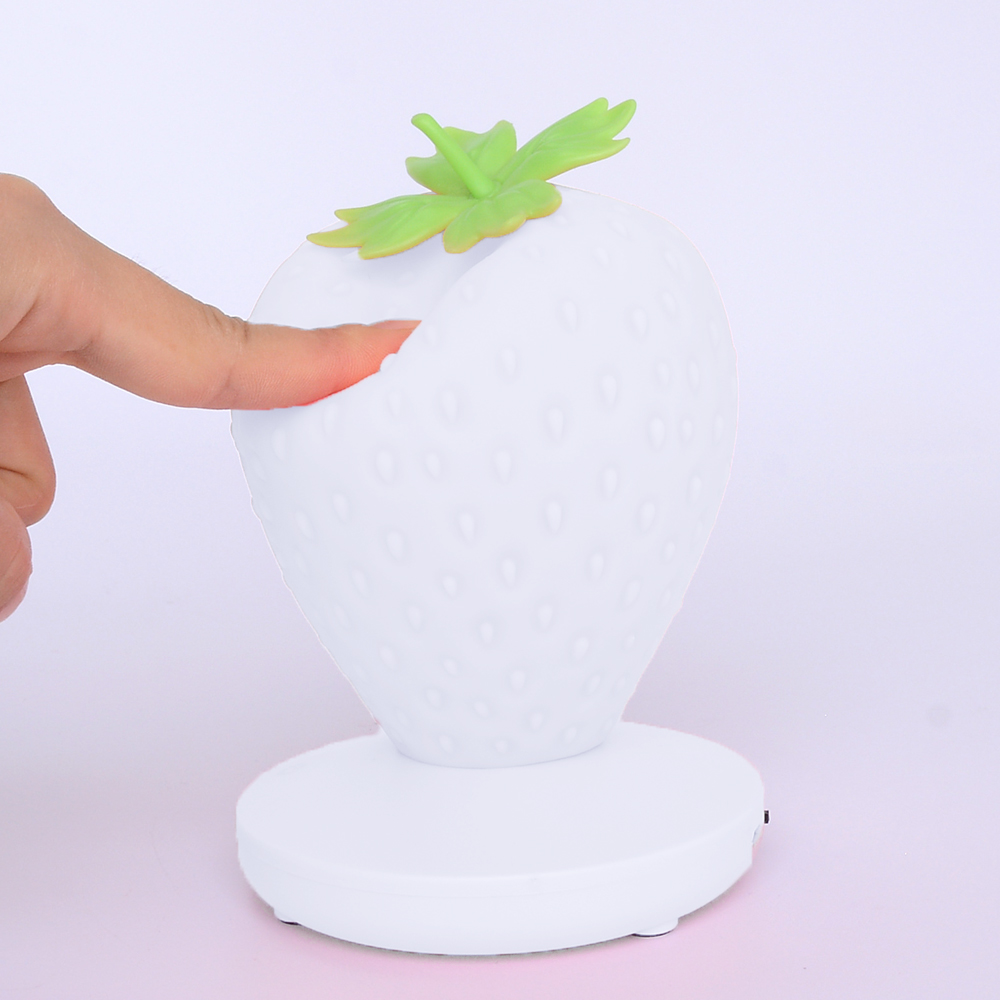 Eine weiße erdbeerförmige Lampe mit grünen Blättern. Ein Finger berührt die Lampe. Die Lampe steht auf einem weißen Sockel, vor rosa Hintergrund.