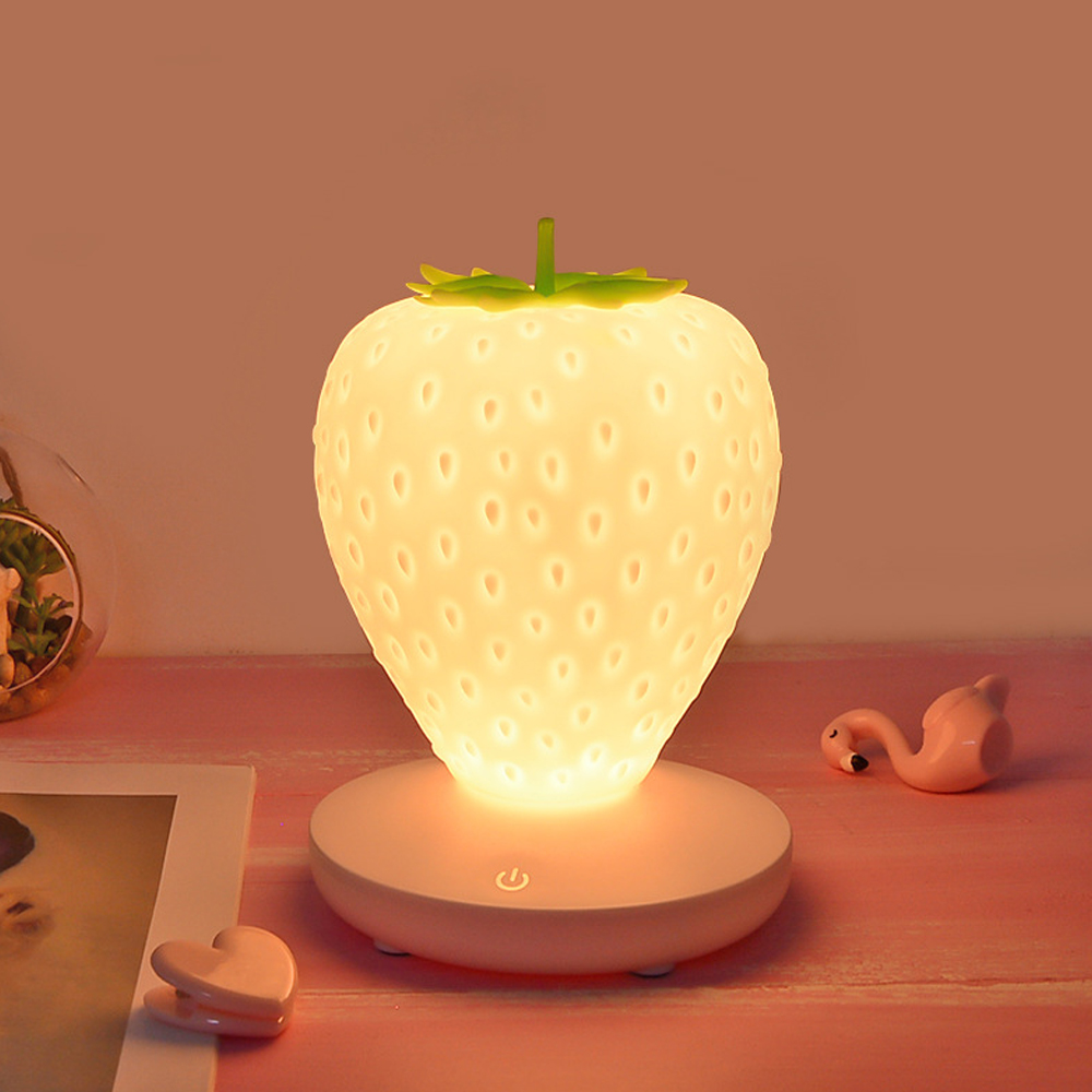 Eine erdbeerförmige Lampe leuchtet. Sie steht auf einem rosa Tisch mit einer Flamingo-Figur und einem herzförmigen Clip. Der Hintergrund ist beige.
