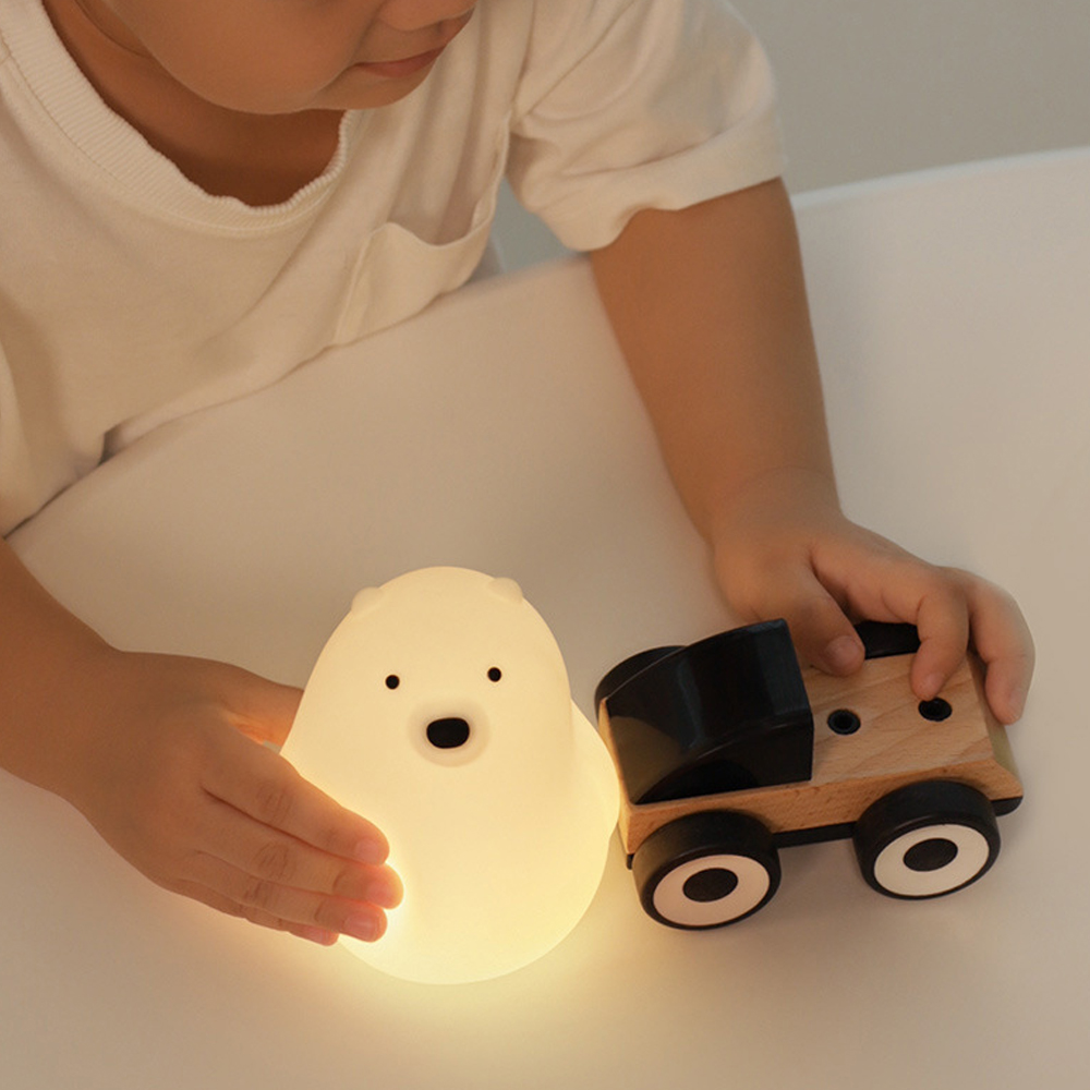 Die Hände eines Kindes berühren eine leuchtende weiße Bärenlampe und einen Holzspielzeug-LKW auf einer weißen Oberfläche.