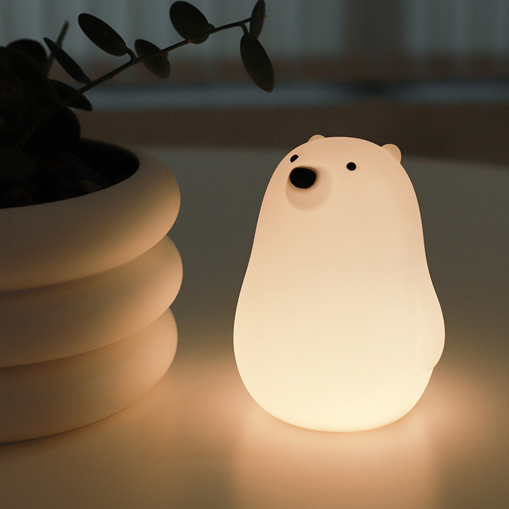 Eine leuchtende, weißbärenförmige Lampe steht auf einem Tisch. Links ist eine Pflanze in einem weißen Topf. Weiches Licht beleuchtet die Szene.