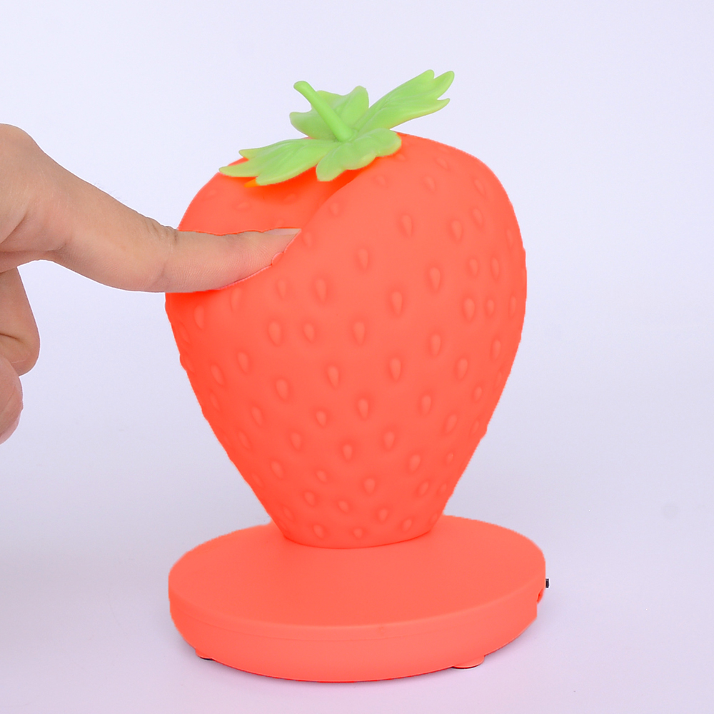 Eine Hand berührt eine erdbeerförmige Lampe. Die Lampe ist orange mit grünen Blättern. Sie steht auf einem runden, orangenen Sockel.
