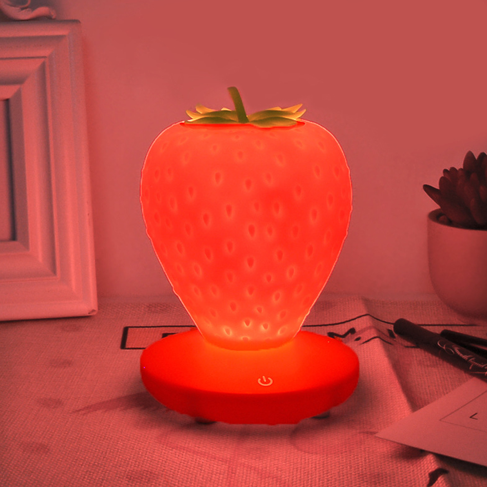 Eine rote erdbeerförmige Lampe leuchtet. Sie steht auf einem roten Sockel. Ein weißer Rahmen und eine Sukkulente sind im Hintergrund.