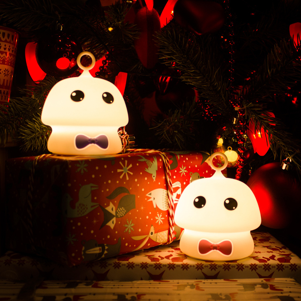 Zwei pilzförmige Lichter mit Fliegen sitzen auf verpackten Geschenken unter einem geschmückten Weihnachtsbaum mit roten Ornamenten.