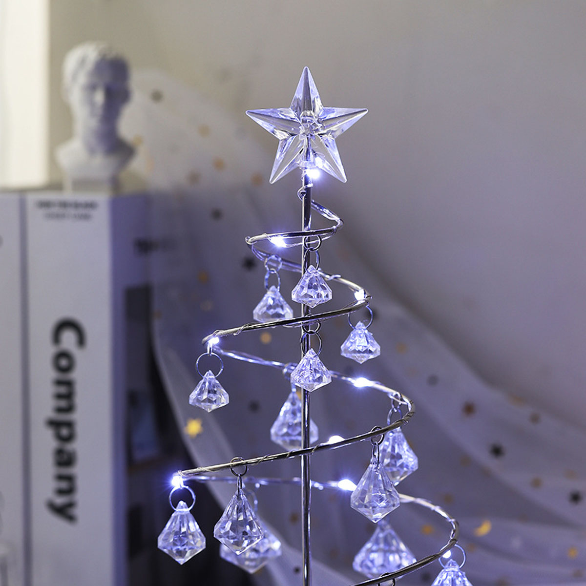 Ein dekorativer silberner Weihnachtsbaum mit Kristallornamenten und einem Stern oben. Im Hintergrund eine Büste und ein Buch.