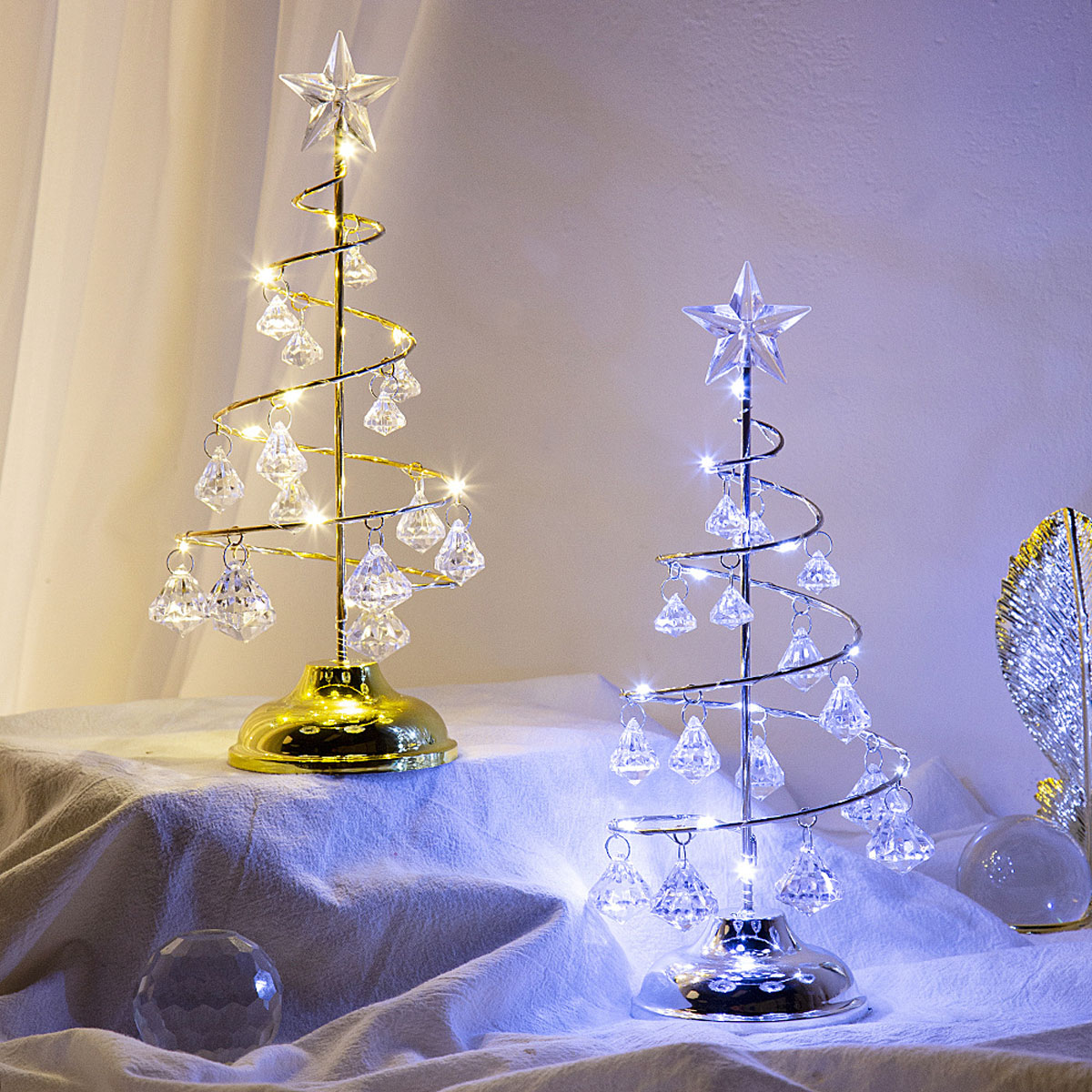 Zwei dekorative Weihnachtsbäume. Einer gold, einer silber, jeweils mit Kristallornamenten und Sternaufsätzen, auf einem weißen Tuch.