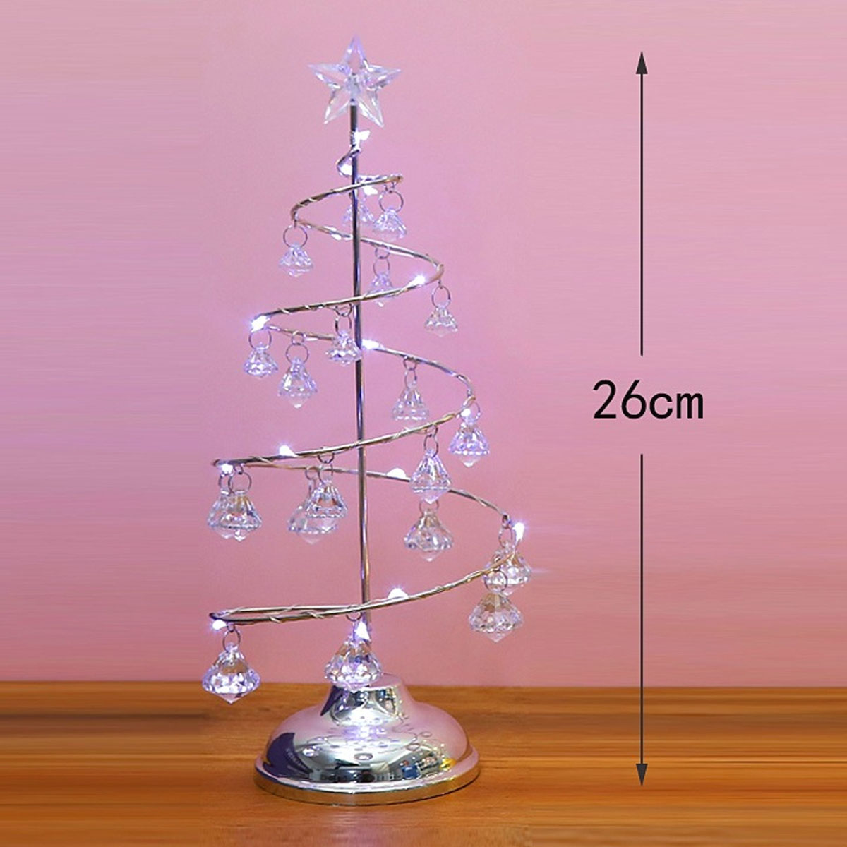 Ein dekorativer silberner Weihnachtsbaum mit hängenden Kristallen und einem Stern. Steht auf einem Tisch vor rosa Hintergrund.