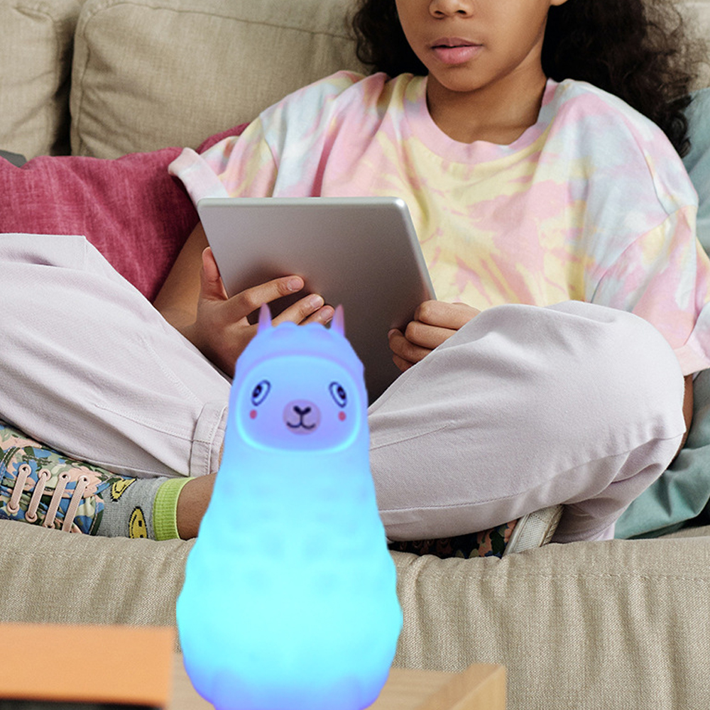 Ein Mädchen sitzt auf einer Couch und hält ein Tablet. Im Vordergrund befindet sich ein blaues, lamaförmiges Nachtlicht.