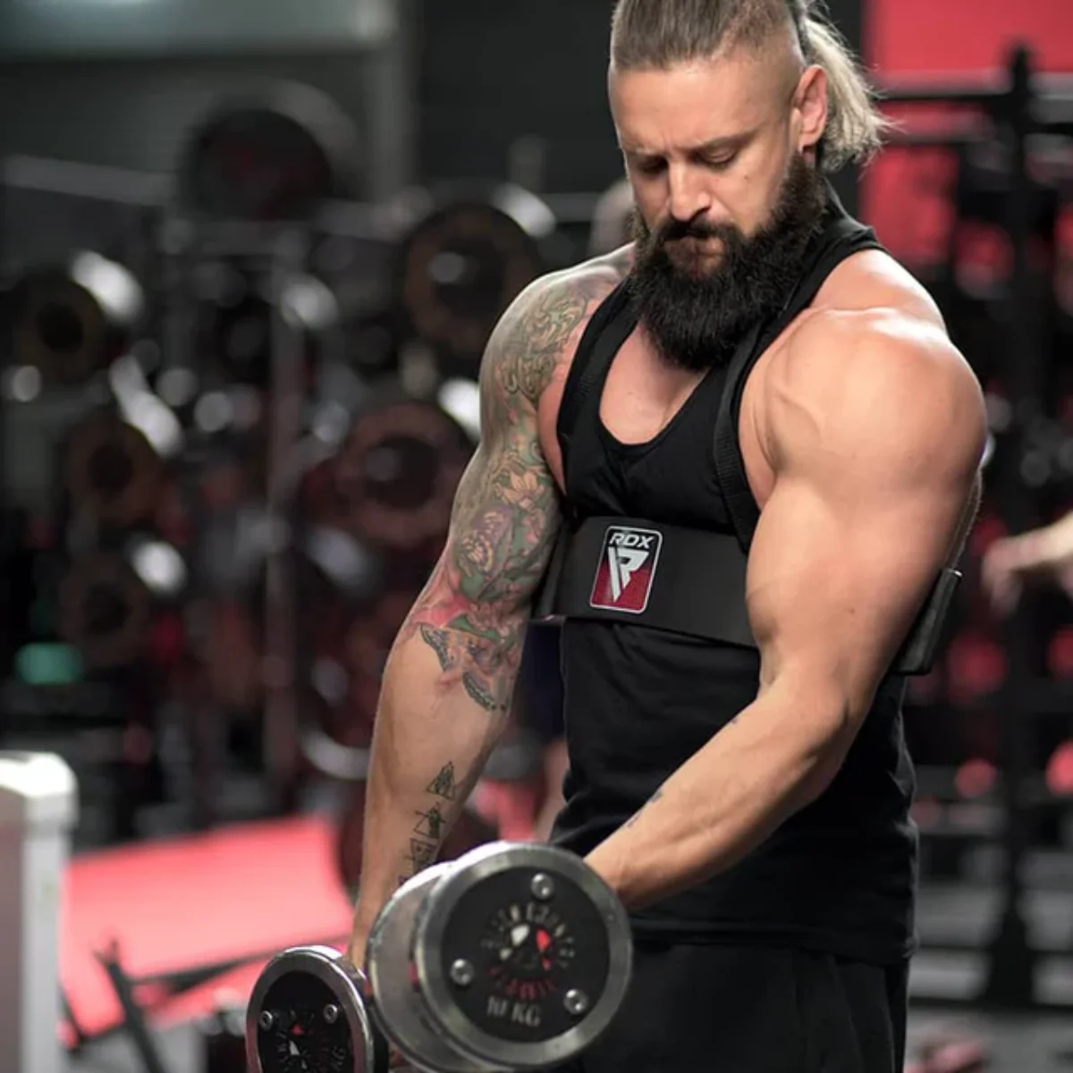 A muscular man with a beard lifts dumbbells in a gym. He wears a black vest and arm support. Tattoos and gym equipment are visible.