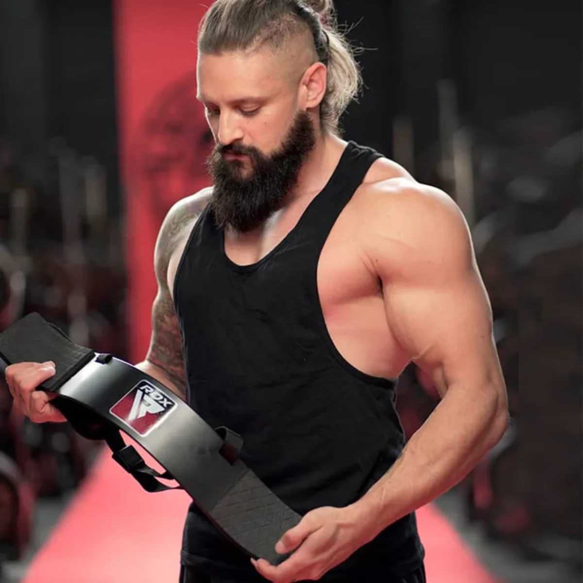 A muscular man with a beard holds a black weightlifting belt. He wears a black tank top in a gym setting. Red background.