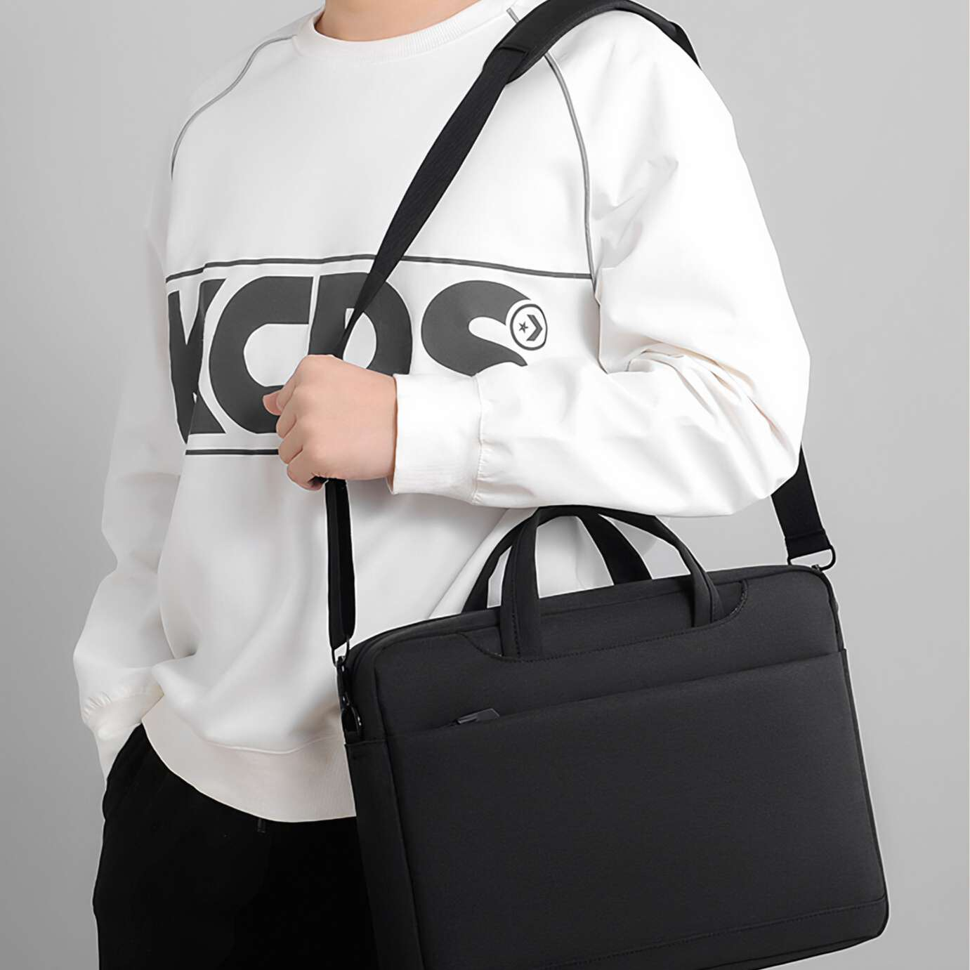 Eine Person hält eine schwarze Laptoptasche. Sie trägt einen weißen Pullover mit schwarzem Text und eine schwarze Hose. Der Hintergrund ist grau.