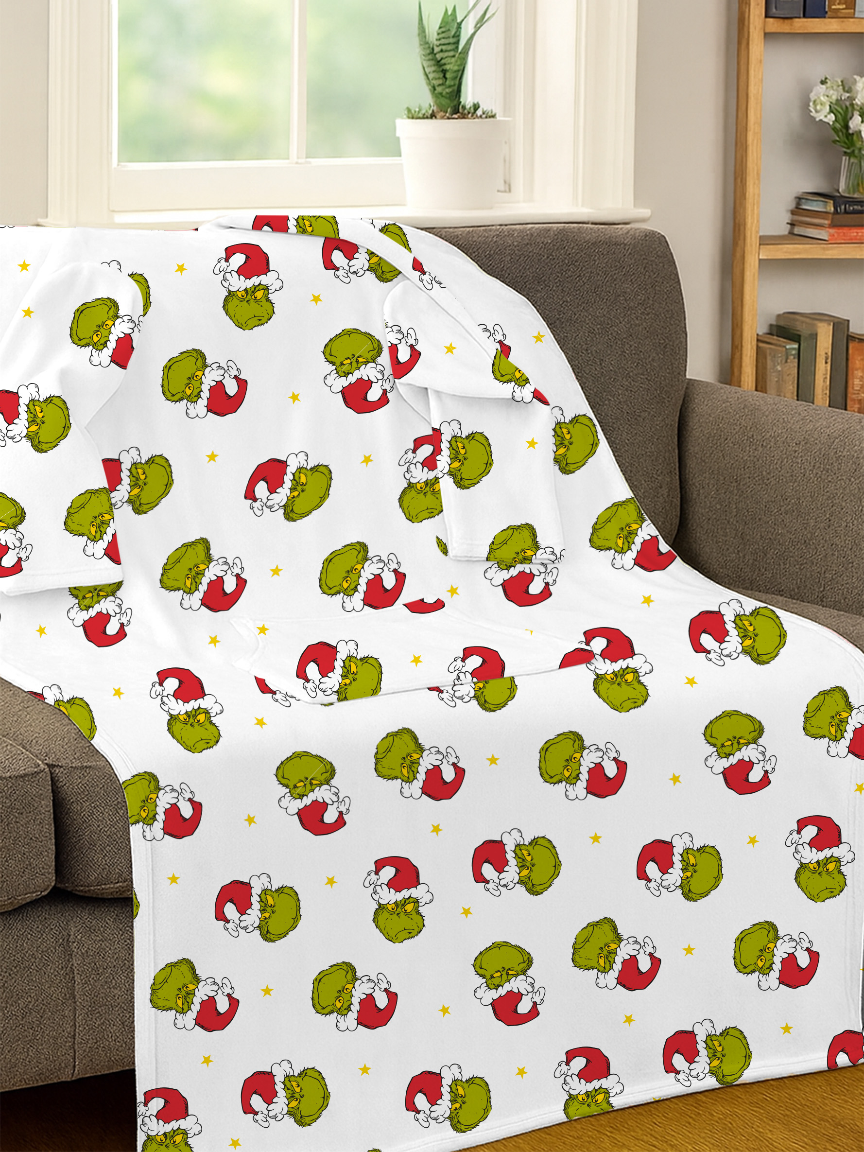 Weiße Decke mit Grinch-Gesichtern in roten Weihnachtsmützen und gelben Sternen auf einem grauen Sofa.