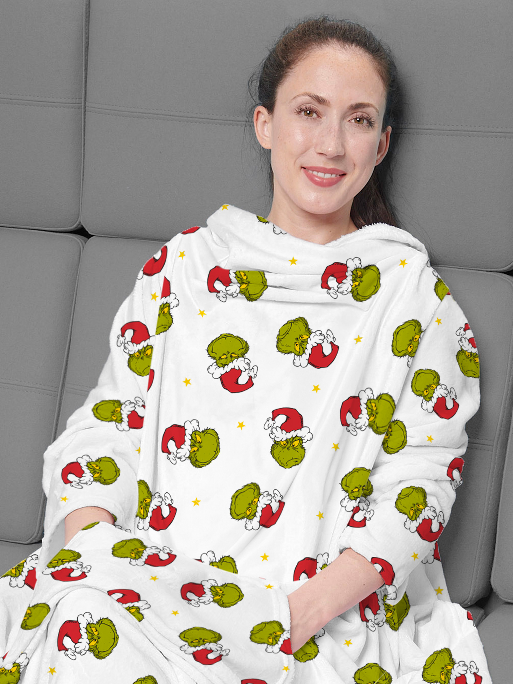 Eine lächelnde Frau trägt eine Grinch-Decke. Die Decke ist weiß mit Grinch-Gesichtern und roten Weihnachtsmützen. Sie sitzt auf einem grauen Sofa.
