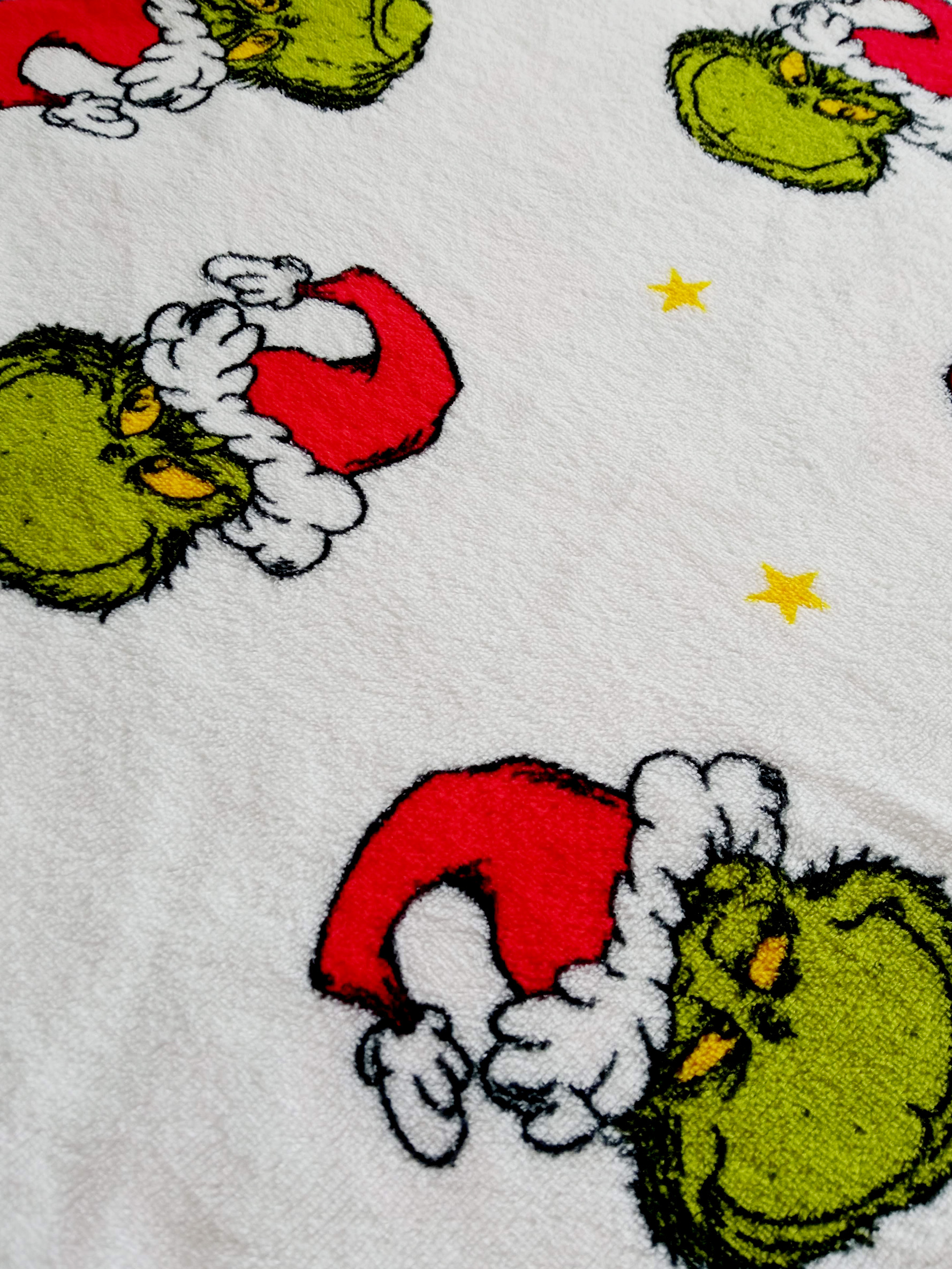 Ein weißes Handtuch mit sich wiederholenden Grinch-Gesichtern, die rote Weihnachtsmützen tragen und gelbe Sterne.