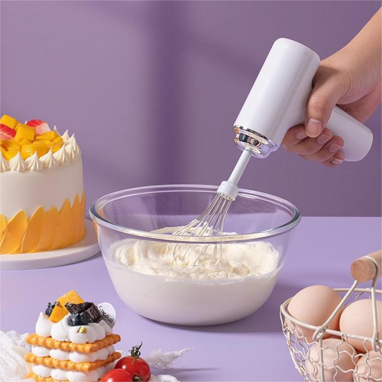 Eine Hand hält einen weißen Mixer über einer Schüssel Sahne. Kuchen, Eier und Tomaten liegen in der Nähe auf einer lila Oberfläche.