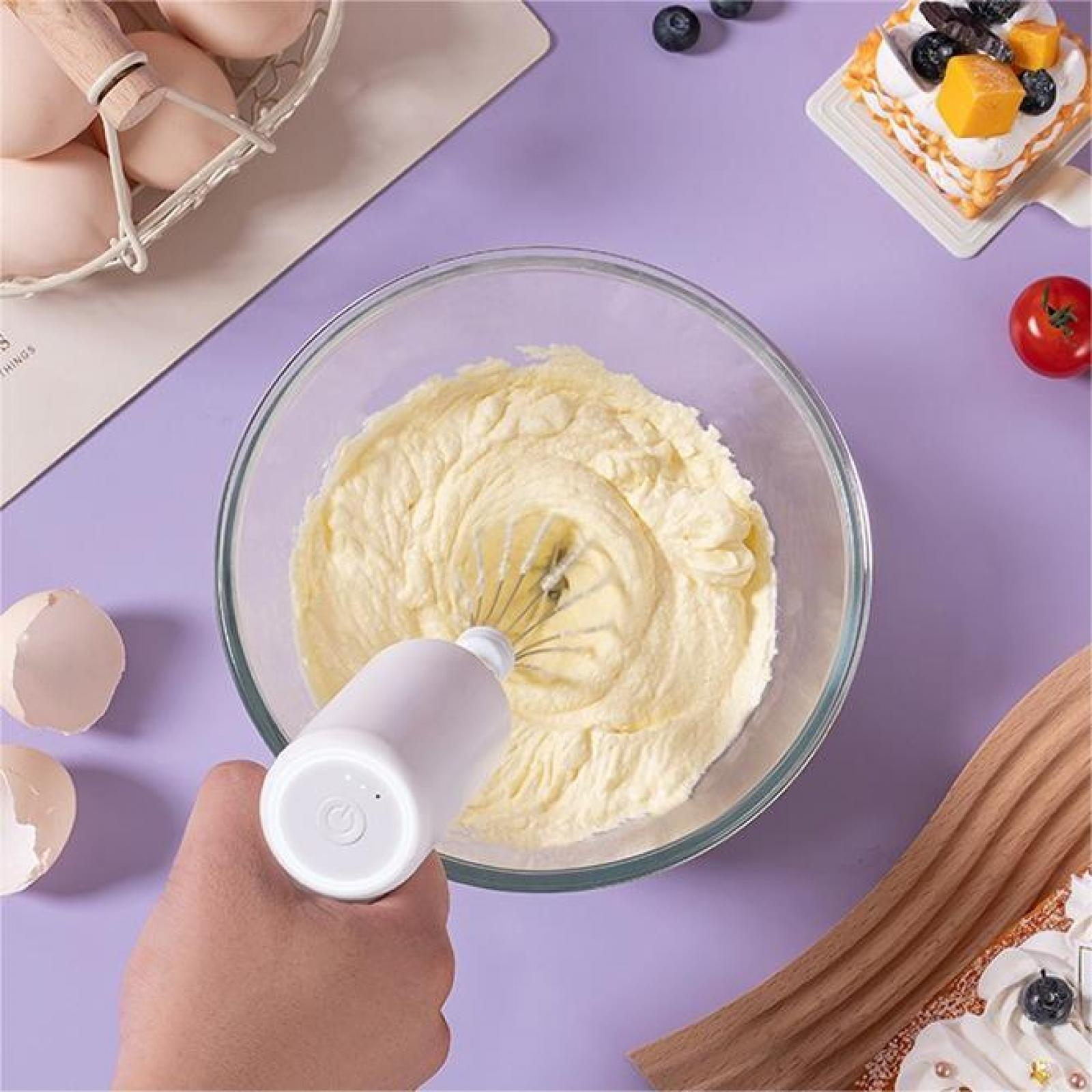 Eine Hand, die einen weißen Mixer in einer Schüssel mit Teig hält. Eier, ein Kuchen und eine Tomate liegen auf einer lila Oberfläche.