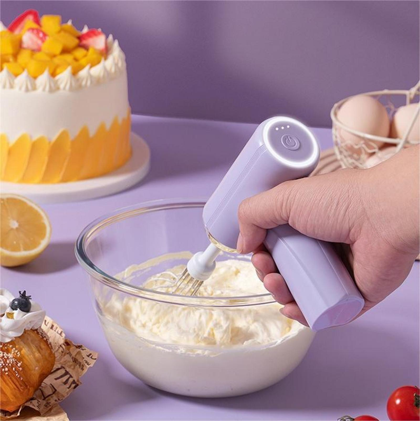 Eine Hand, die einen lila Mixer hält und Sahne in einer Schüssel verrührt, mit Kuchen, Eiern und Gebäck im Hintergrund.