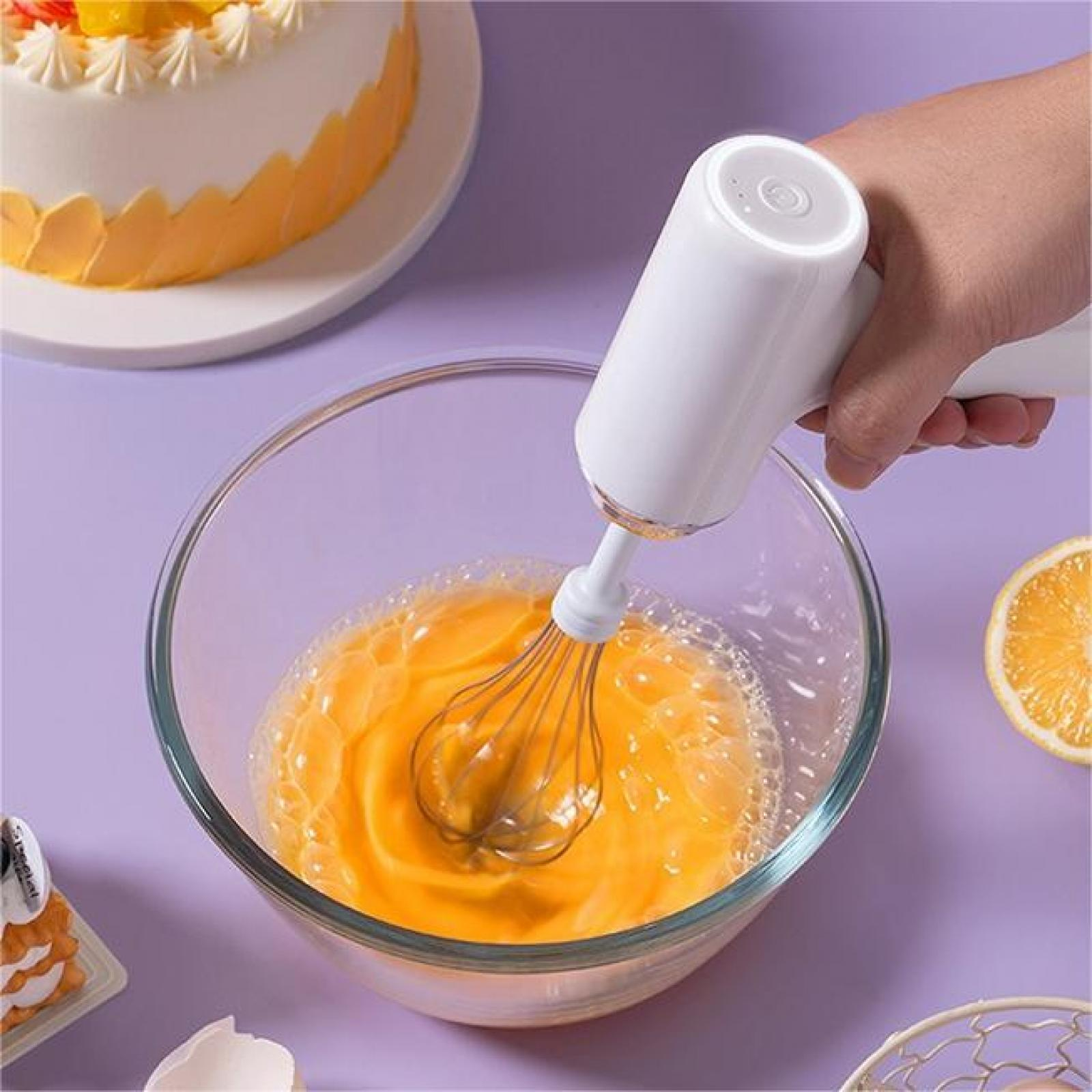 Eine Hand, die einen weißen elektrischen Mixer hält und Eier in einer Glasschüssel verrührt. Ein Kuchen und eine Orangenscheibe sind im Hintergrund.