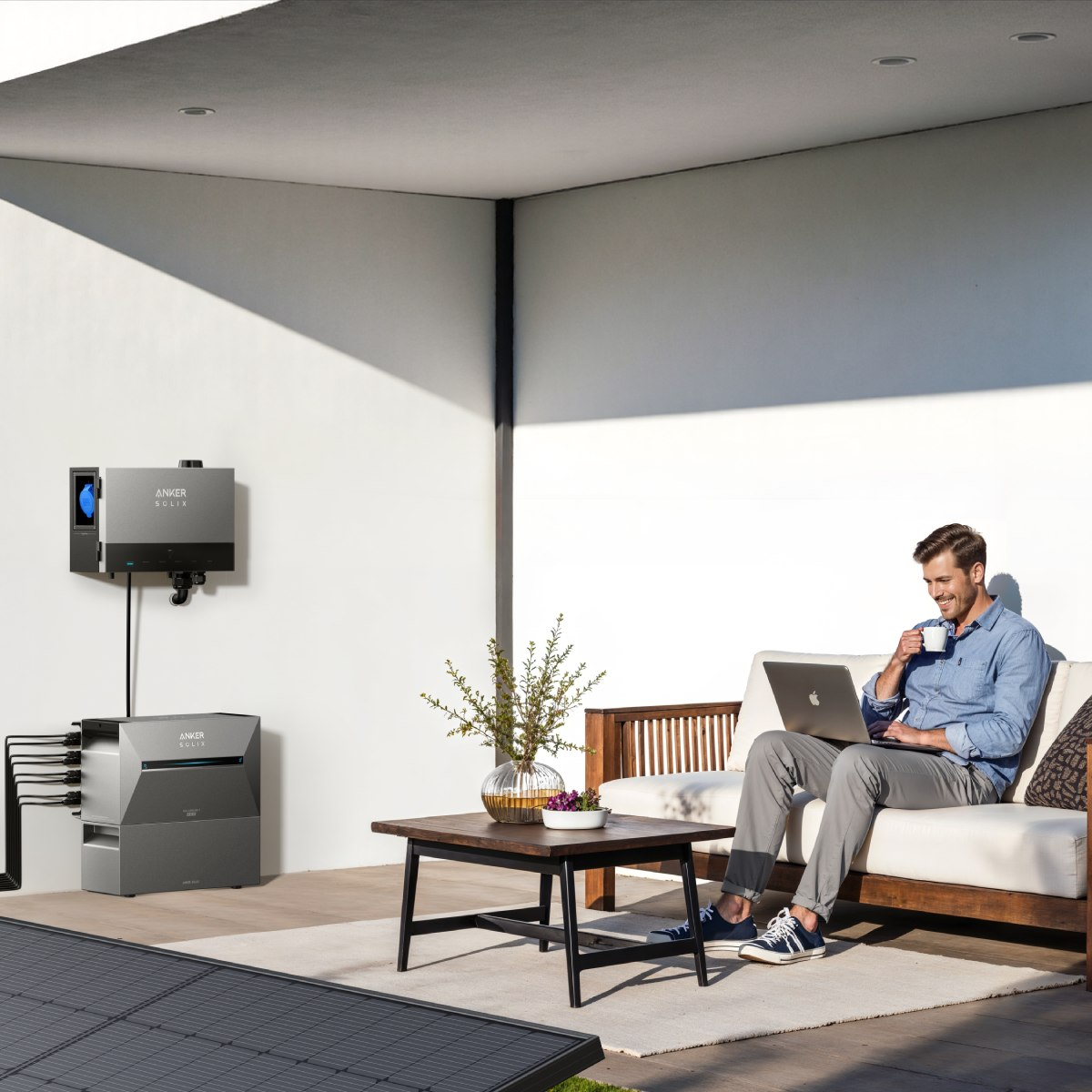 Ein Mann sitzt draußen, benutzt einen Laptop und trinkt Kaffee. Im Vordergrund befinden sich Sonnenkollektoren und ein Energiespeichersystem.