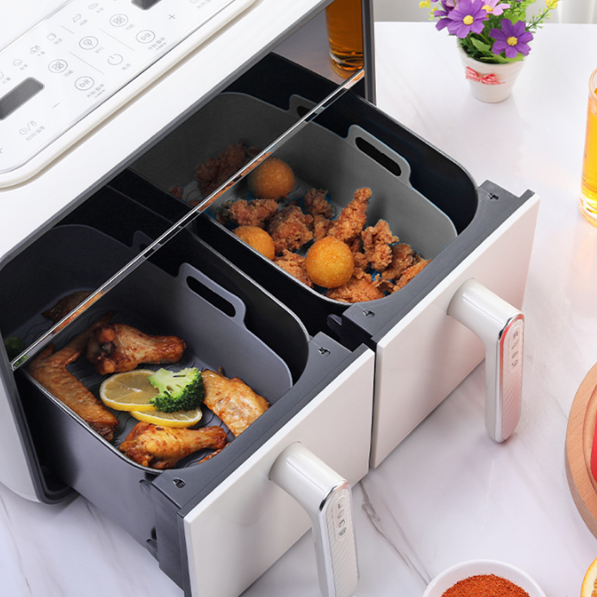 Eine Heißluftfritteuse mit Hähnchenflügeln, Frittiertem und Zitronenscheiben in Fächern. Ein Blumentopf und ein Glas Saft sind in der Nähe.