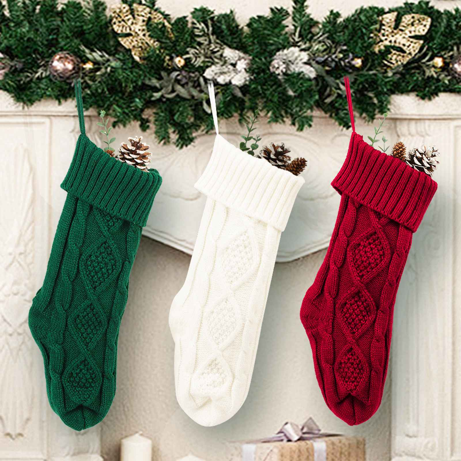 Drei gestrickte Weihnachtsstrümpfe: grün, weiß und rot, gefüllt mit Tannenzapfen und Grünzeug, hängen über einem Kamin.