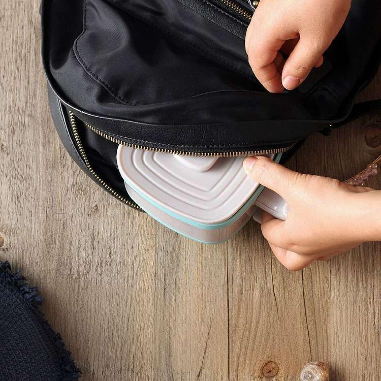 Eine Person legt eine Brotdose auf einer Holzoberfläche in einen schwarzen Rucksack.