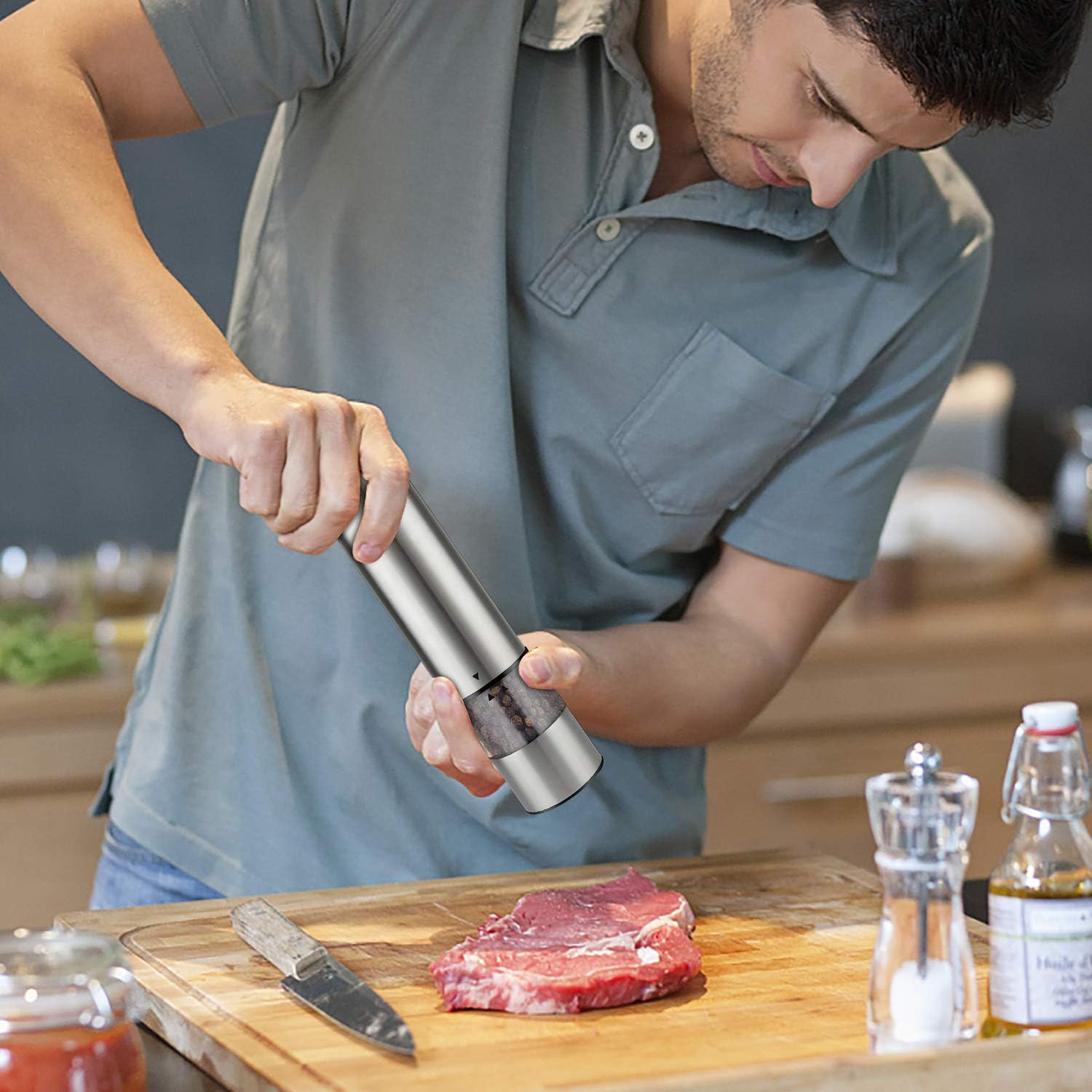 Ein Mann mahlt Pfeffer auf rohes Fleisch auf einem Holzbrett. Ein Messer und Ölflaschen sind in der Nähe. Er trägt ein graues Hemd.