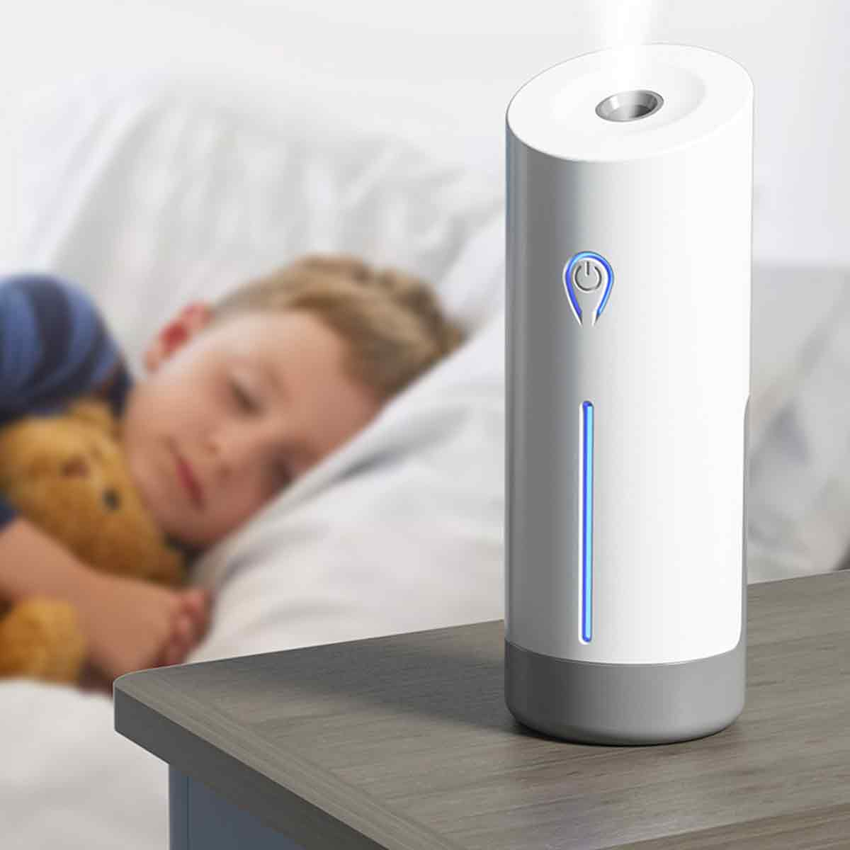 Ein weißer Diffusor steht auf einem Nachttisch. Im Hintergrund schläft ein Kind, das einen Teddybären umarmt. Der Diffusor hat ein blaues Licht.