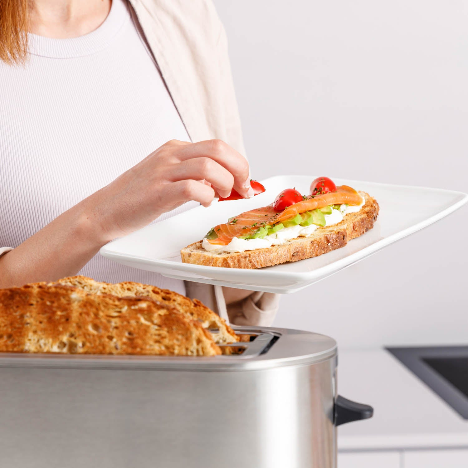 Una persona sta guarnendo un toast con pomodoro. Il toast ha crema, avocado e salmone. Un tostapane è in primo piano.