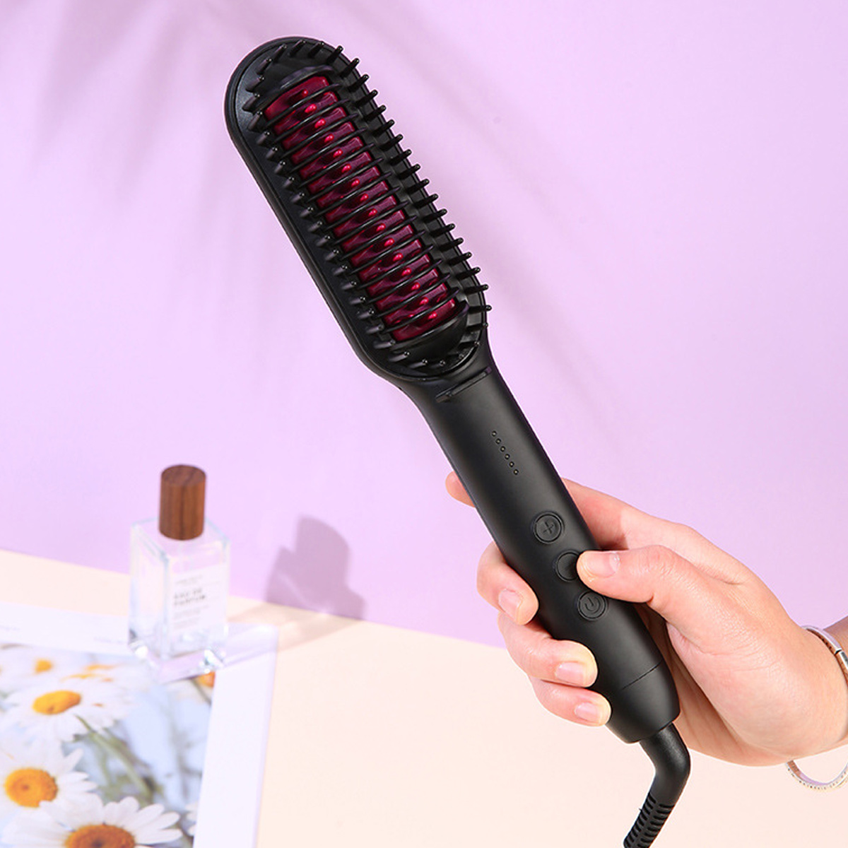 Eine schwarze Haarglättungsbürste wird von einer Hand gehalten. Eine Parfümflasche und ein Blatt mit Blumen sind links.