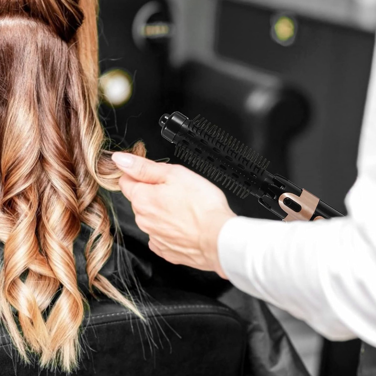 Eine Person benutzt eine Lockenbürste auf welligem, blondem Haar. Die Bürste ist schwarz und goldfarben. Der Hintergrund ist unscharf.