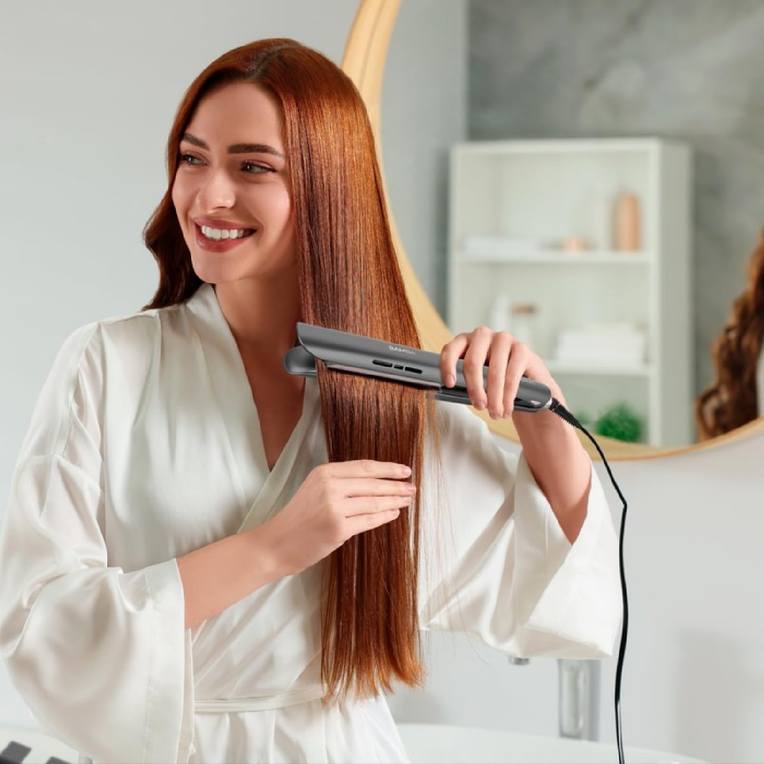 A woman with red hair straightens her hair with a flat iron in a bathroom, wearing a white robe and smiling at the mirror.
